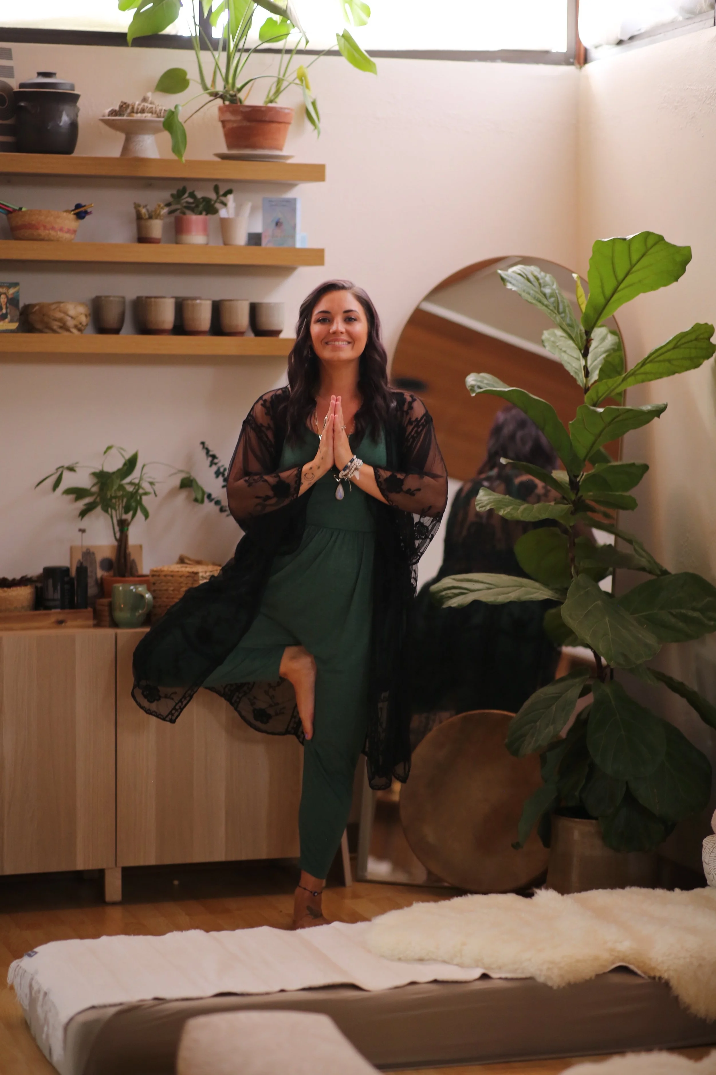 A woman practicing yoga in a cozy, decorated room, standing on one leg with hands in prayer position, smiling, surrounded by plants and wooden furniture.