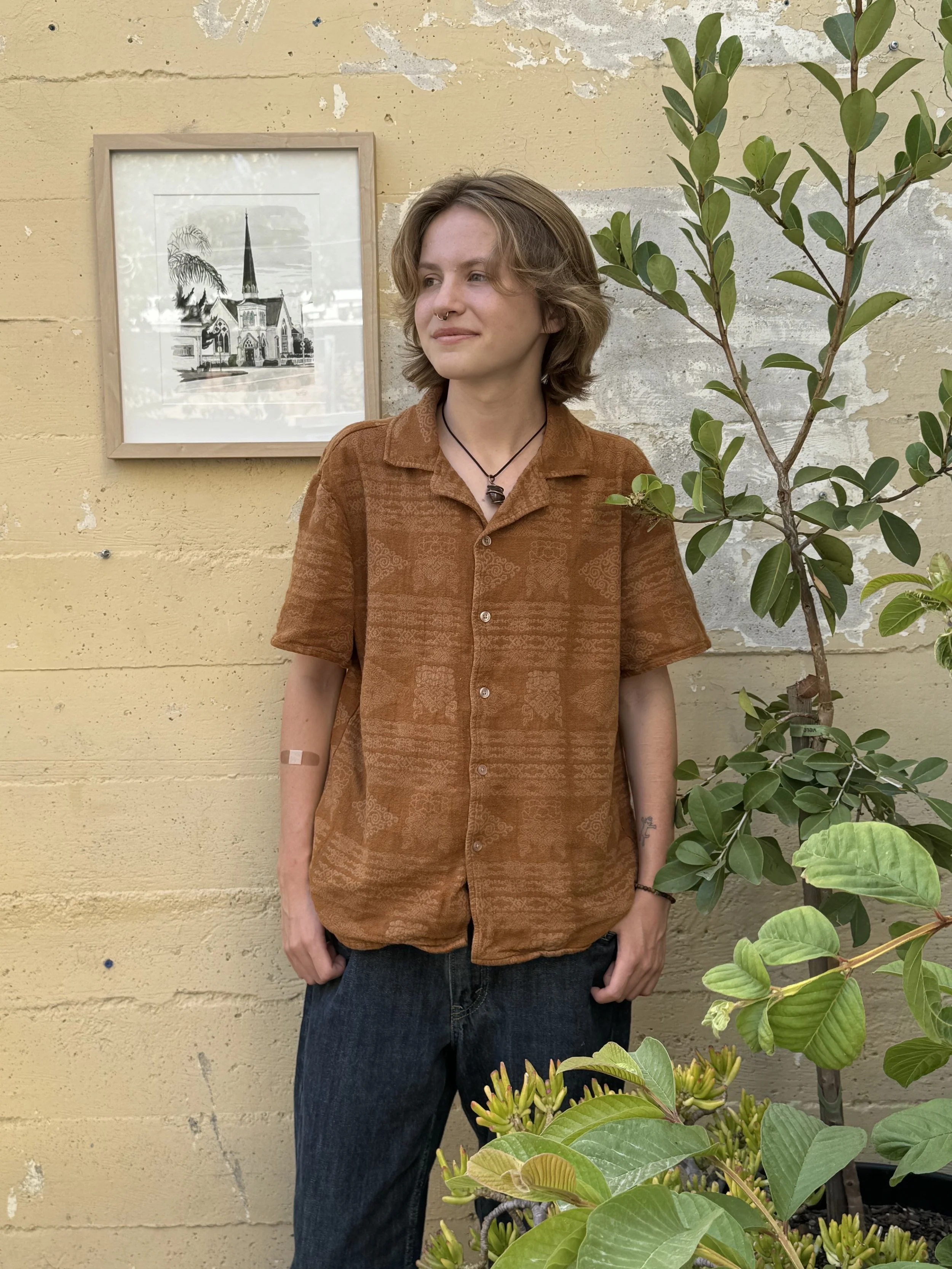 A young person with shoulder-length brown hair, wearing a brown patterned short-sleeve shirt and dark jeans, stands outdoors next to a green leafy plant and a yellow brick wall. There is a framed black and white drawing of a church with a tall steeple behind them.