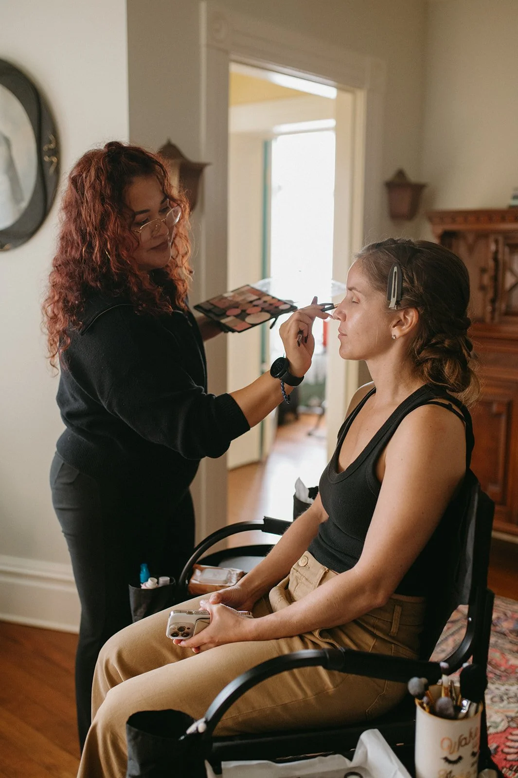 Makeup artist applying eye makeup to a woman sitting in a wheelchair, holding a phone, in a cozy room with wooden furniture and a doorway in the background.