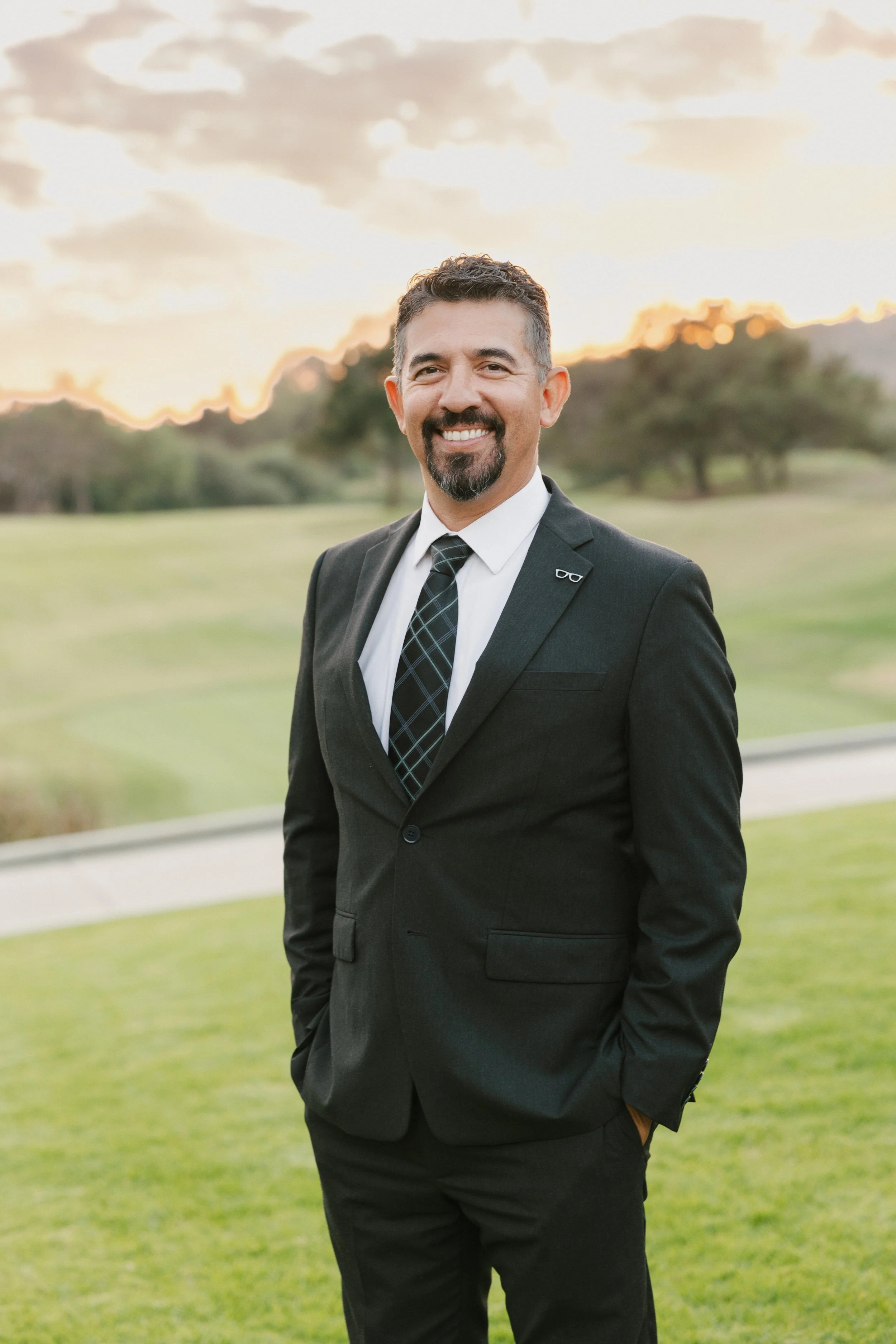 A man in a black suit, white shirt, and plaid tie smiling outdoors at sunset, with trees and green grass in the background.