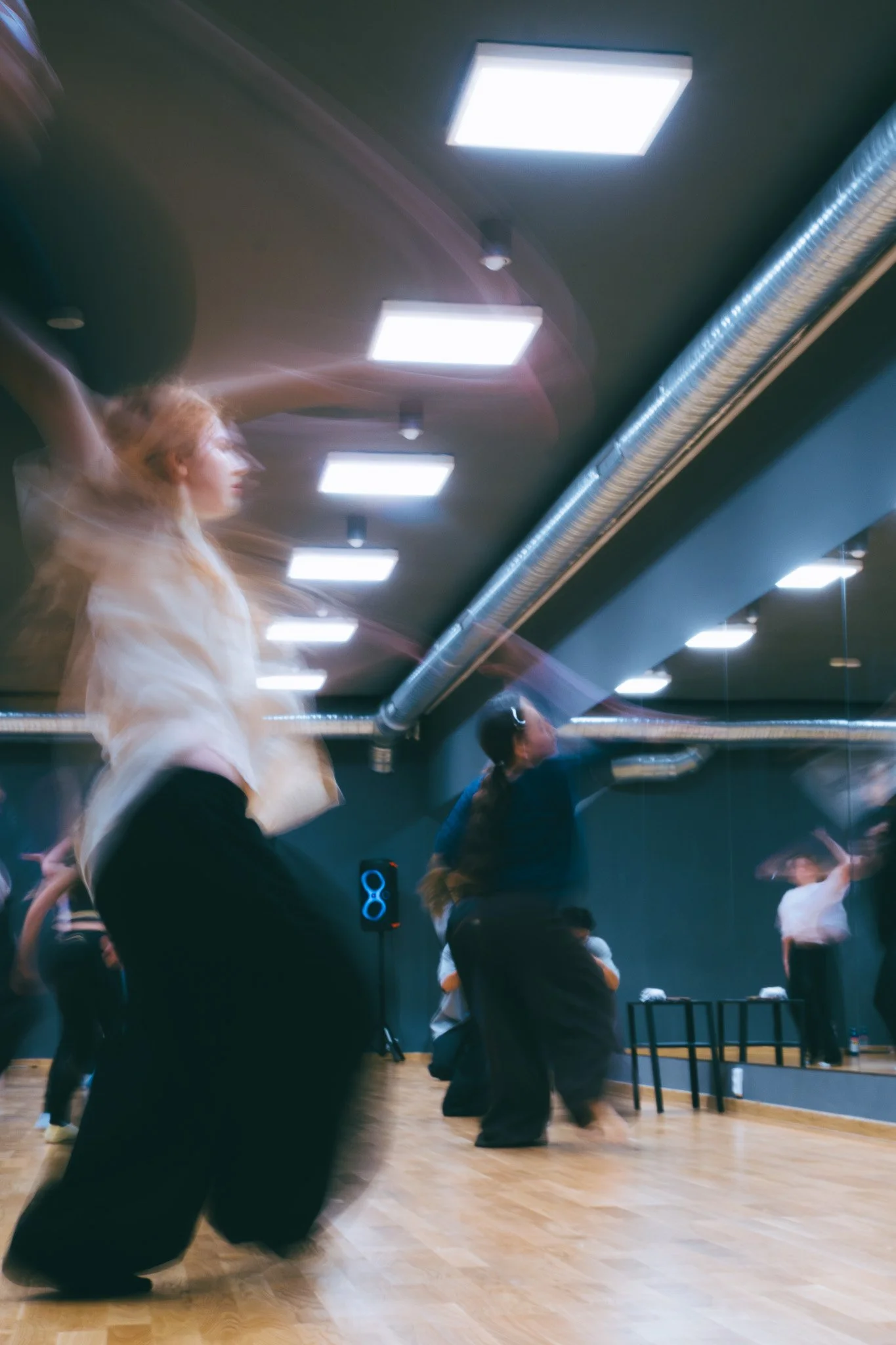 Dance class with students practicing ballet in a studio, with a woman in a white top and black pants in the foreground and others dancing behind her.