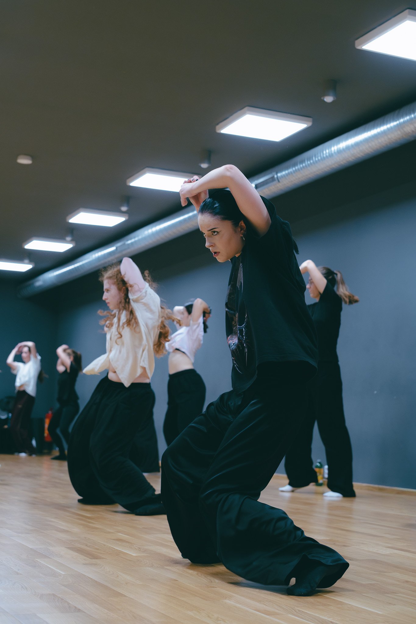 A group of women practicing contemporary dance in a studio with wooden floors and ceiling lights, performing a synchronized move with arms raised and body bent.
