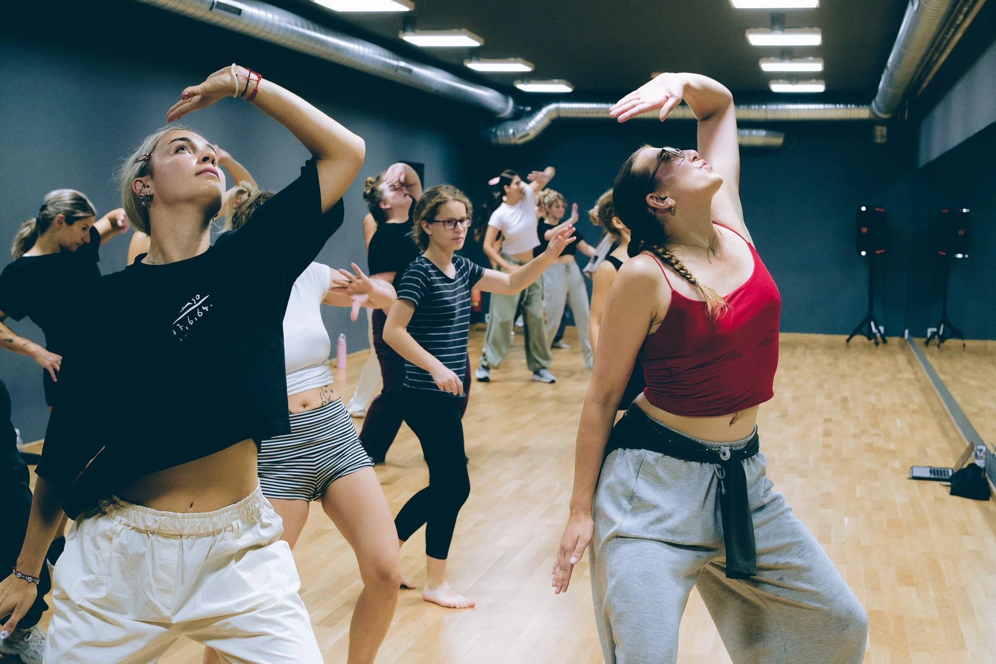 People participating in a dance or exercise class in a studio with wood flooring and gray walls, some in casual wear and others in workout clothes, with their arms raised and in motion.