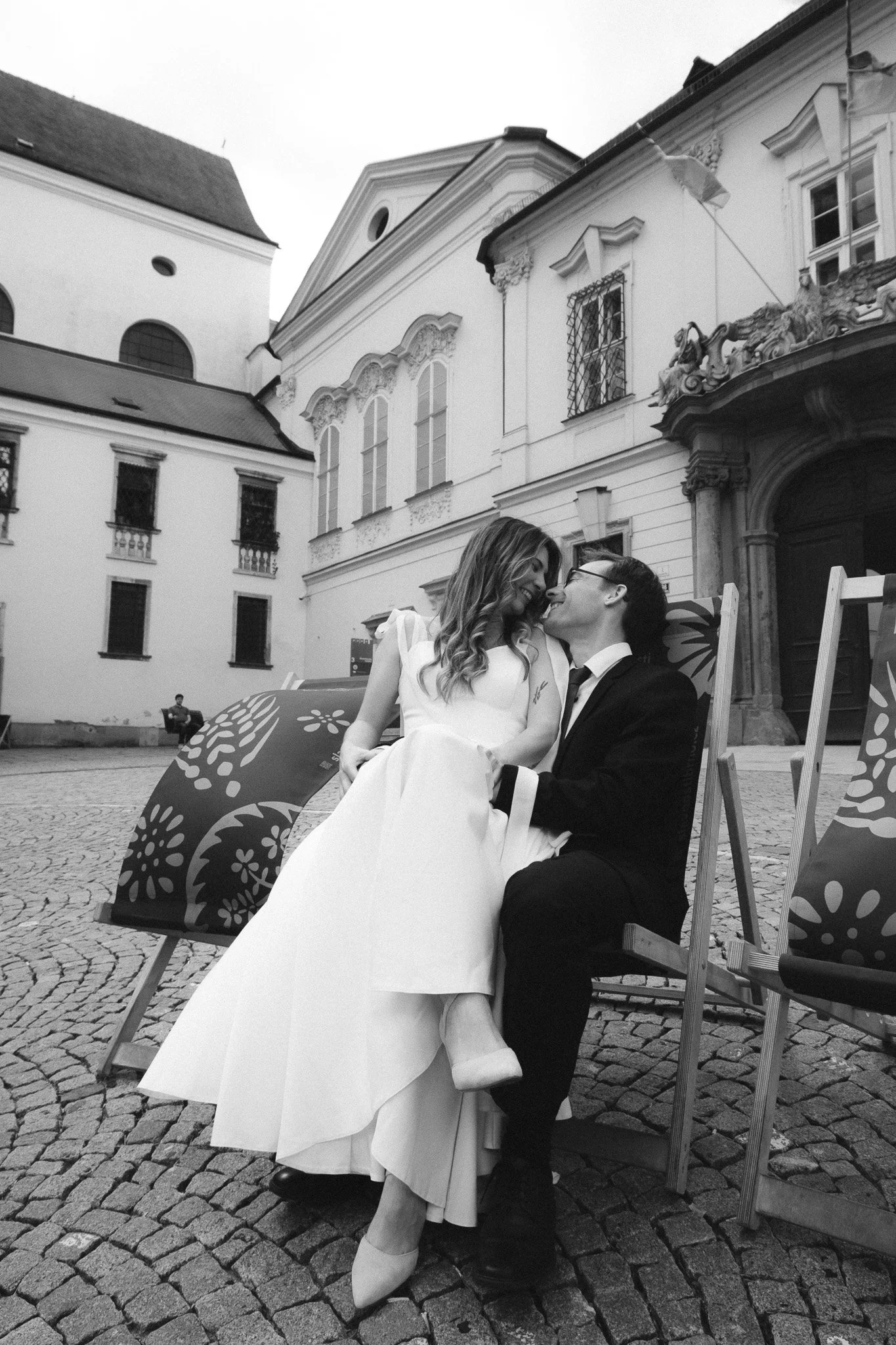 A couple sitting close together outdoors, in front of historic European-style buildings. The woman is wearing a white dress or coat, and the man is in a dark suit with glasses. They are smiling and leaning in towards each other, sharing a joyful moment.