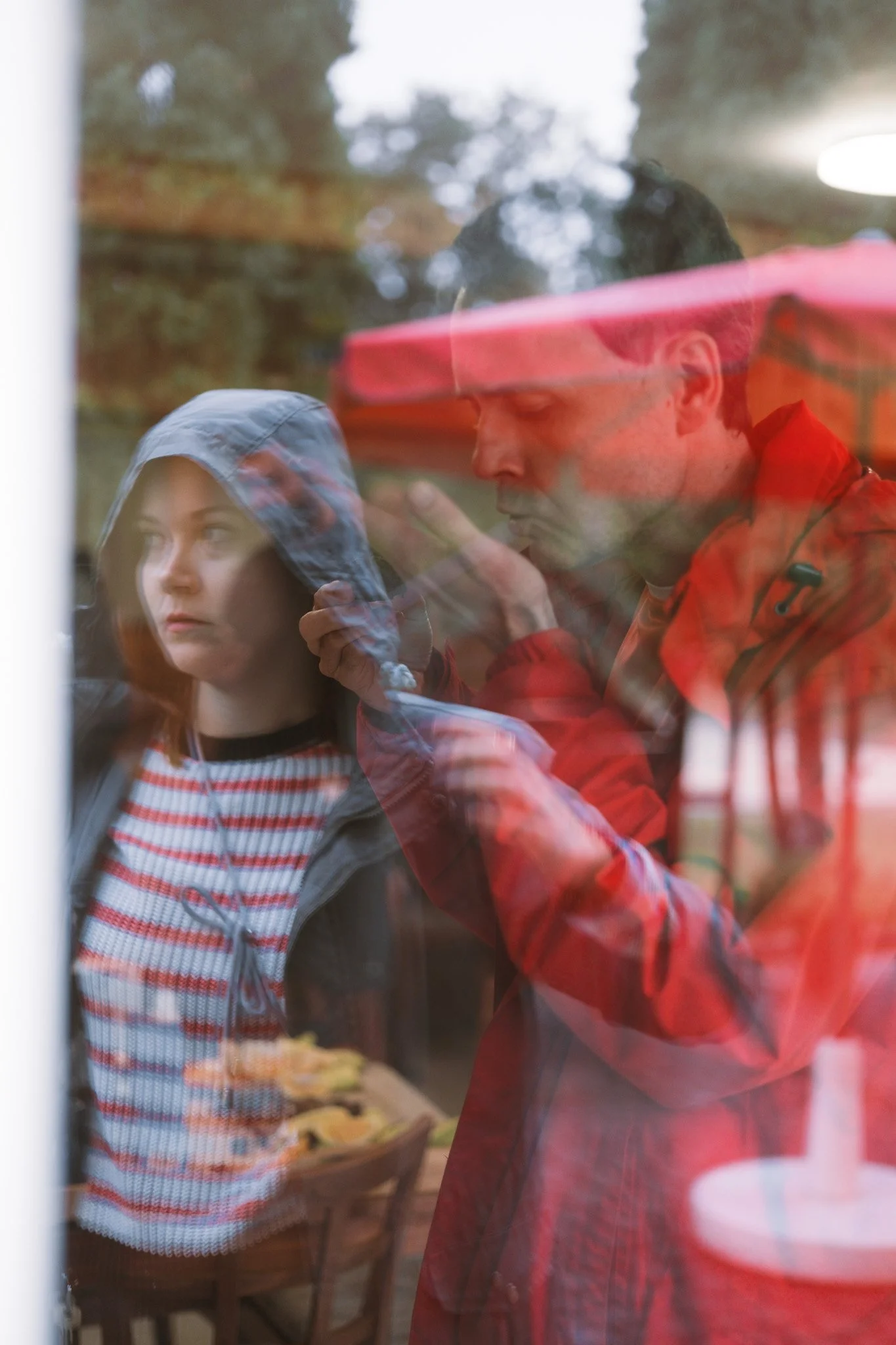 Two people are seen through a glass window. One is a young girl with straight hair wearing a wool hoodie, and the other is an adult man in a red jacket, possibly with glasses, looking down at something or thinking.