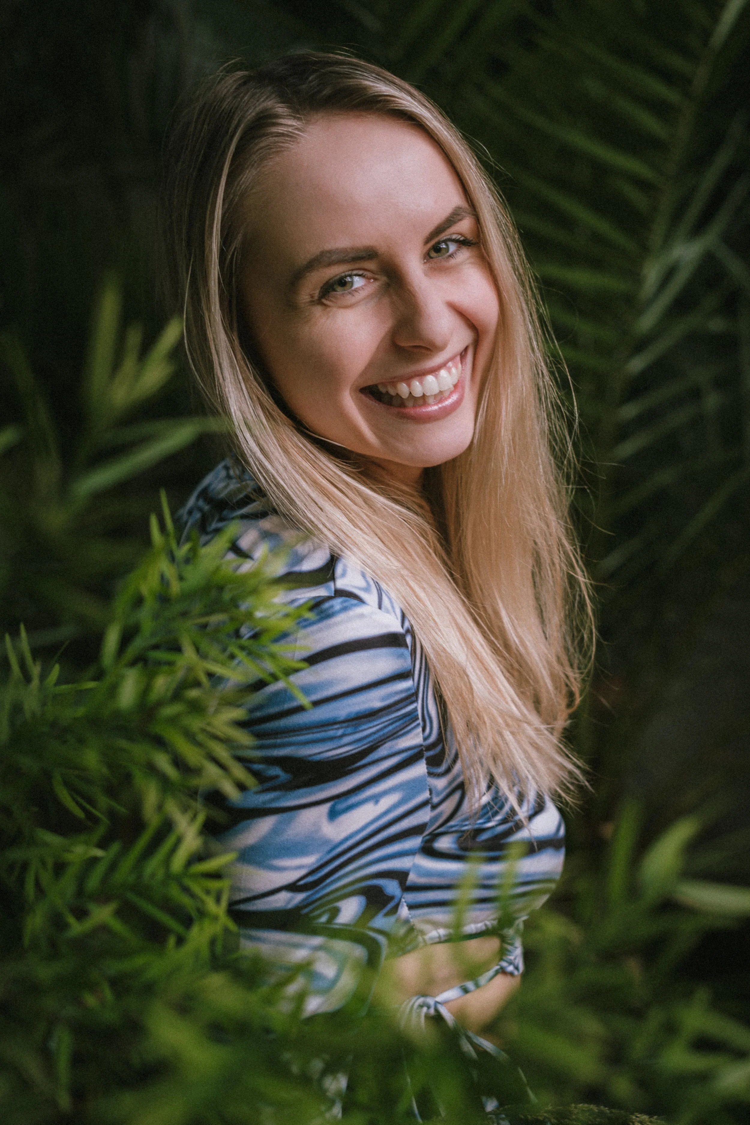 Mladá žena s dlouhými blond vlasy s úsměvem a při pohledu na fotoaparát, stojící mezi zelenými tropickými rostlinami.