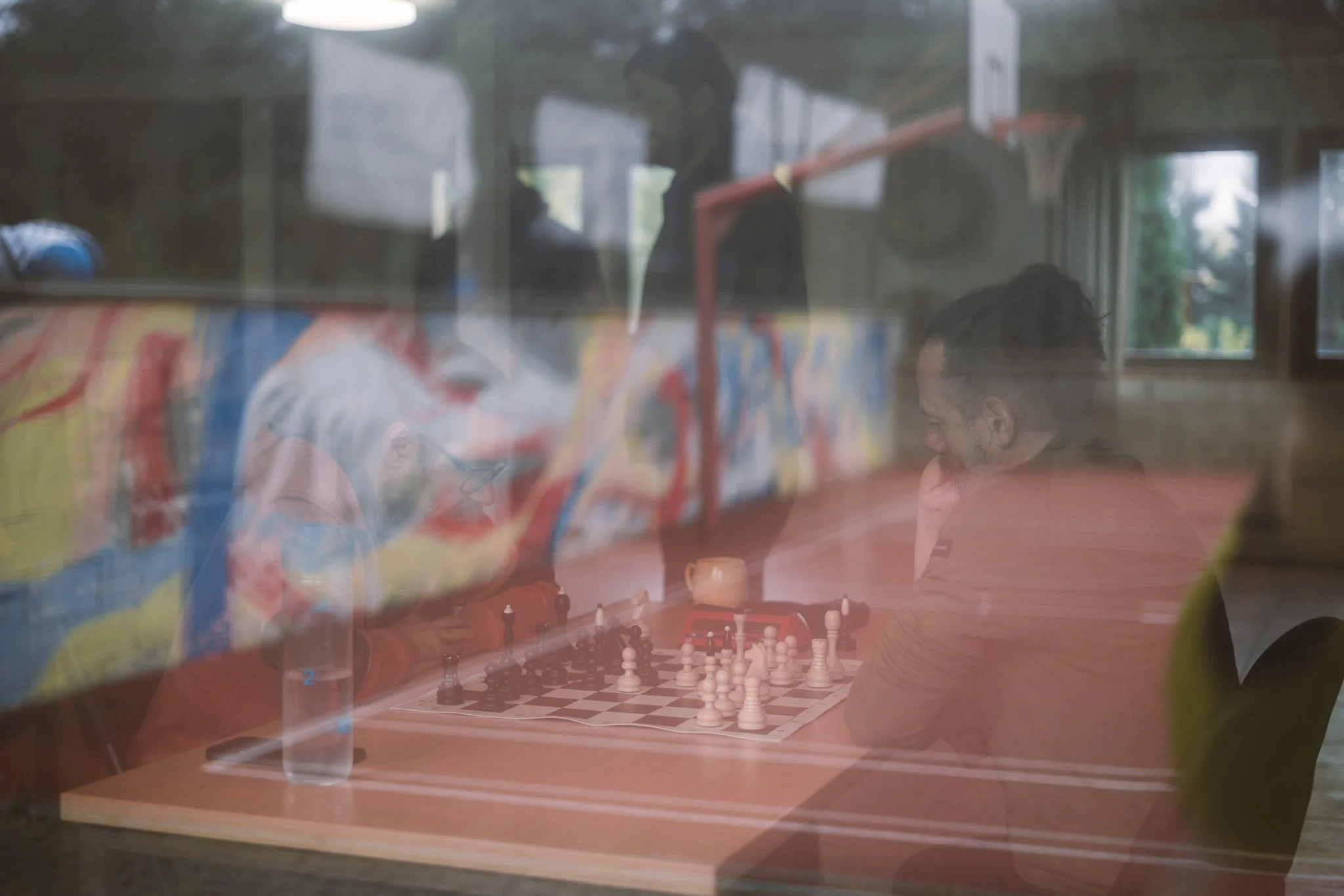 A reflection of children playing chess inside a room with large windows, seen through a glass or plastic surface, creating a layered, blurry effect.