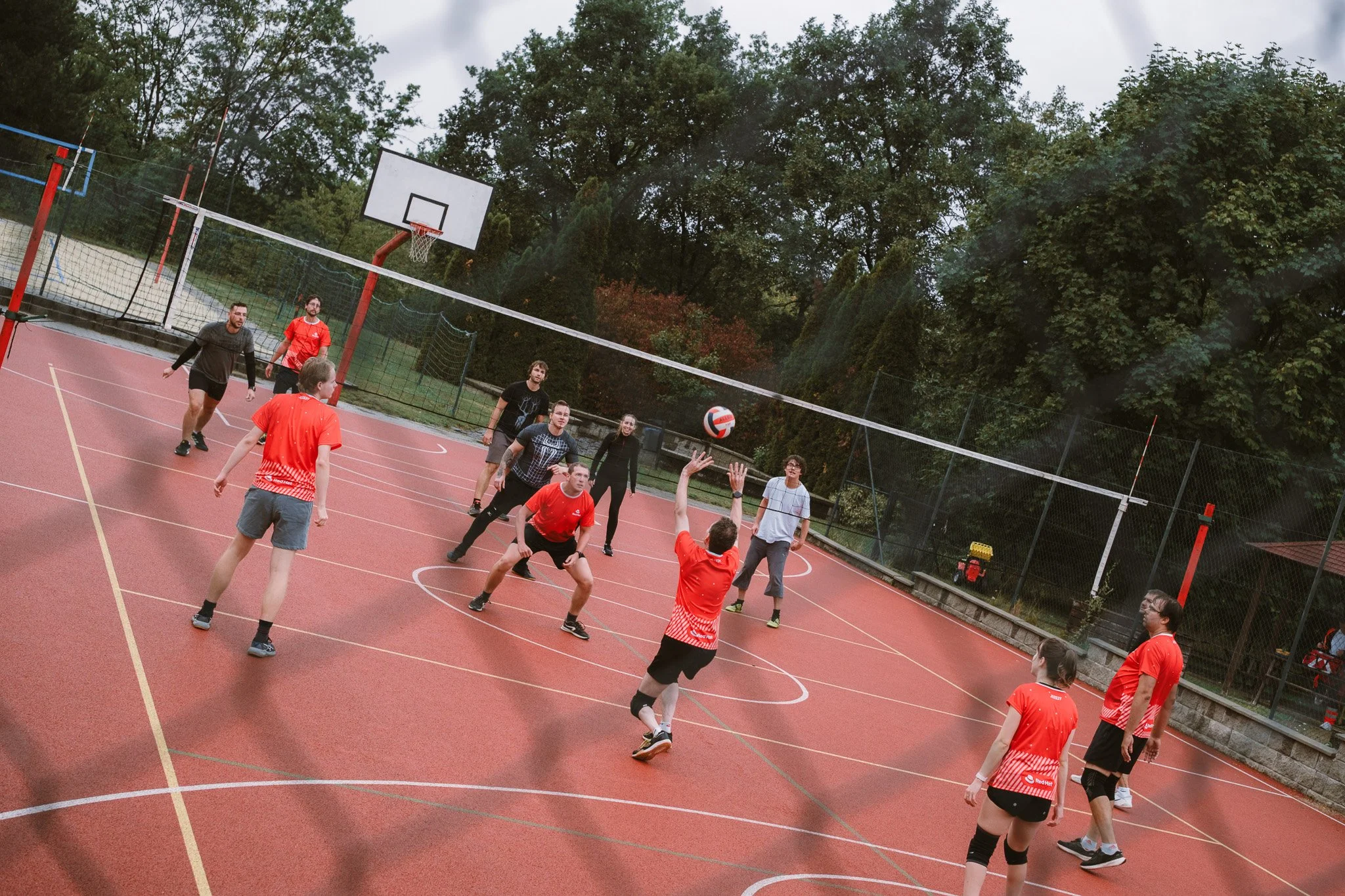 People playing volleyball on an outdoor court surrounded by trees, with a net and a basketball hoop visible.