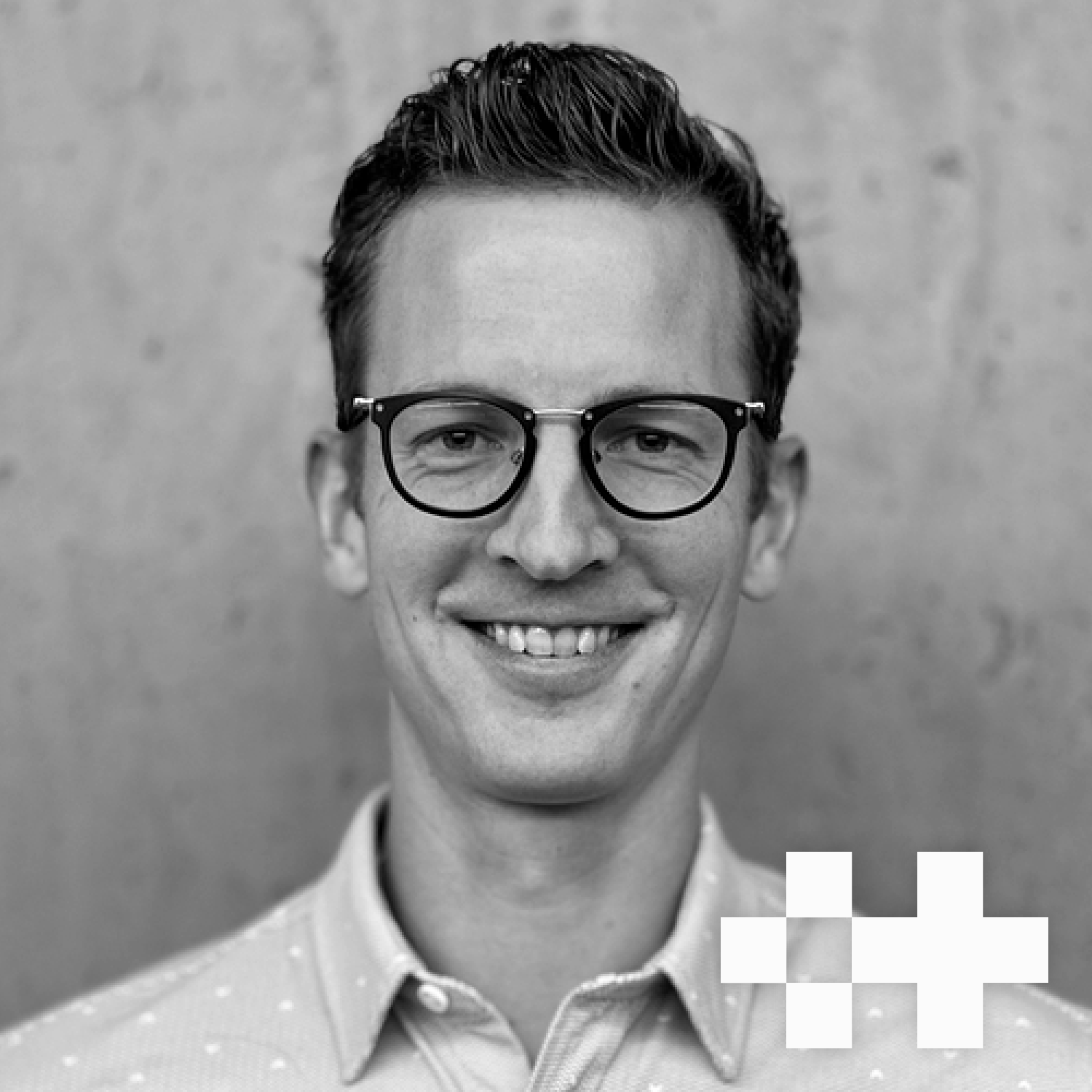 Black and white portrait of a young man with glasses, smiling, wearing a collared shirt with a dotted pattern, standing against a plain wall background.