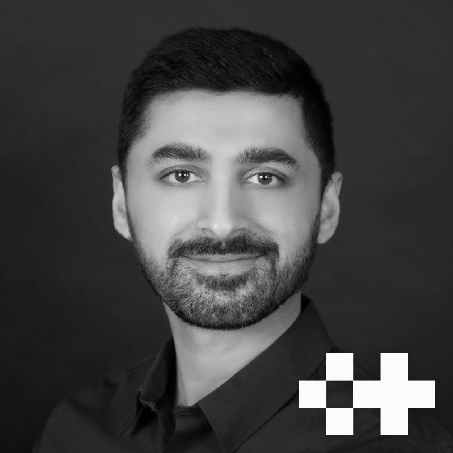 Black and white portrait of a man with short dark hair, beard, and mustache, wearing a dark shirt, smiling softly against a dark background.