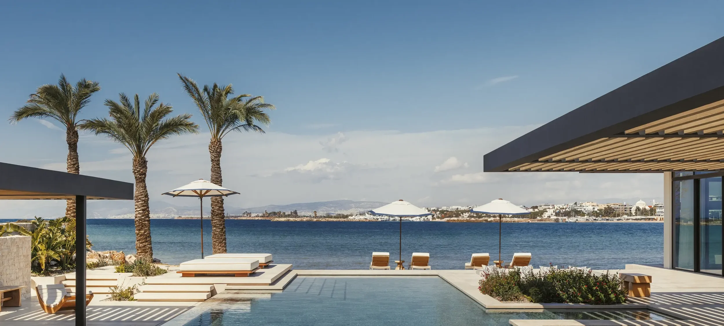 Modern beachfront resort with a pool, sun loungers, umbrellas, and palm trees overlooking the ocean, with a cityscape in the distance under a partly cloudy sky.