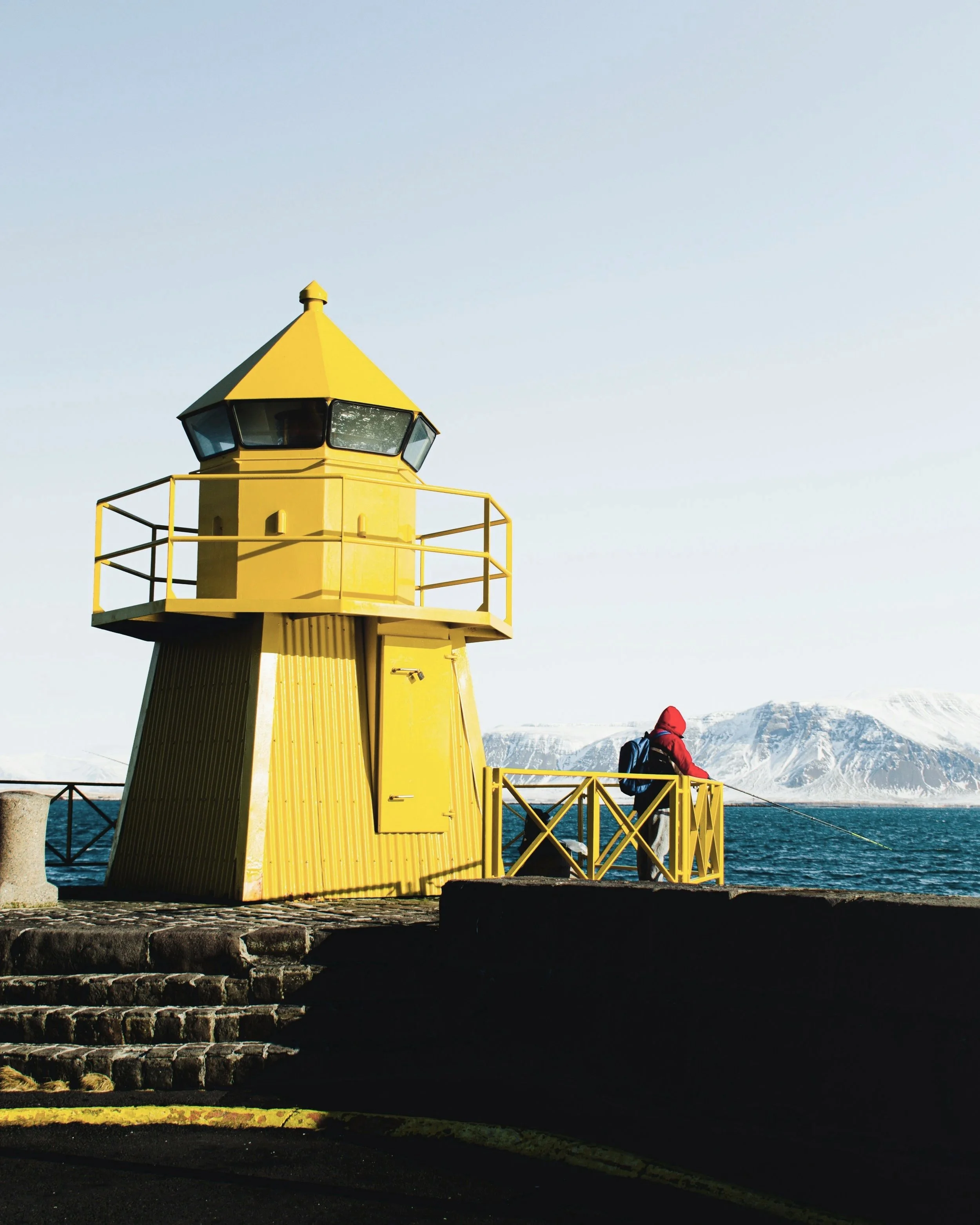 A yellow lighthouse by the sea with a person in a red jacket fishing nearby, snow-covered mountains in the background.