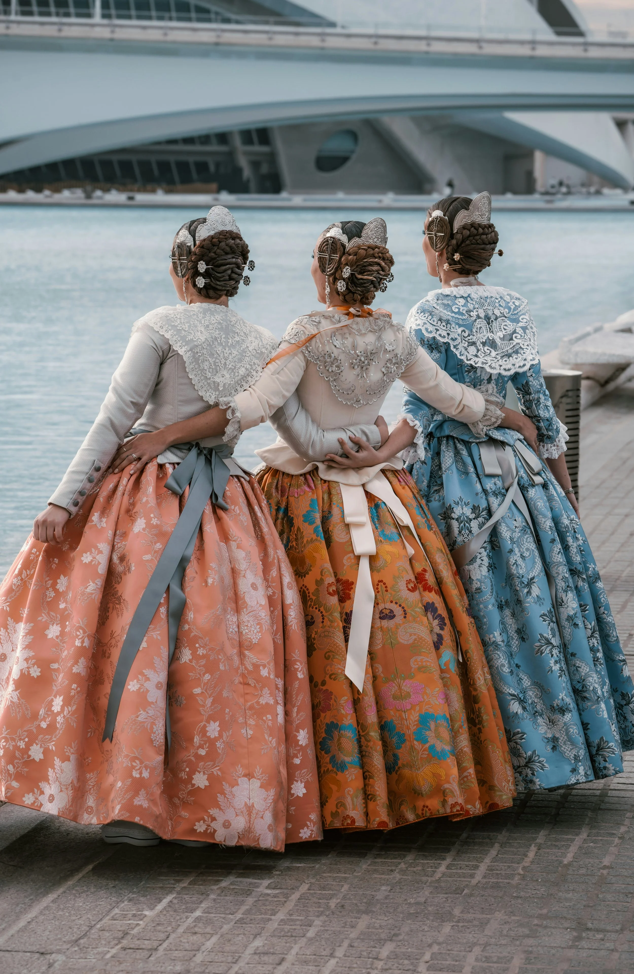 Three women dressed in traditional colorful, floral-patterned, 18th-century style dresses with lace and ruffles, walking along a waterway with modern bridges in the background, holding each other by the waist.