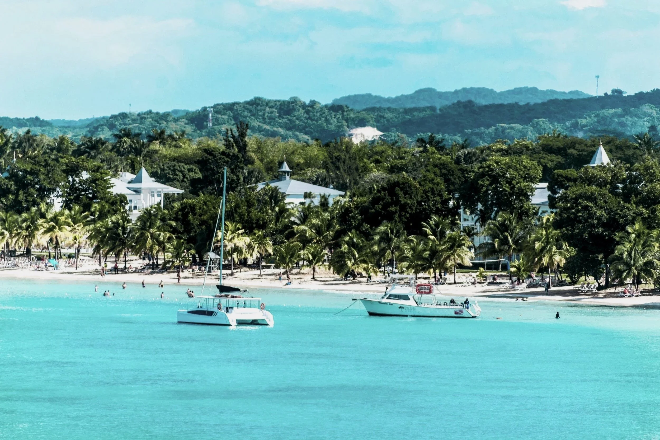 Tropical beach with turquoise water, boats, palm trees, and white buildings in the background, surrounded by lush green hills.