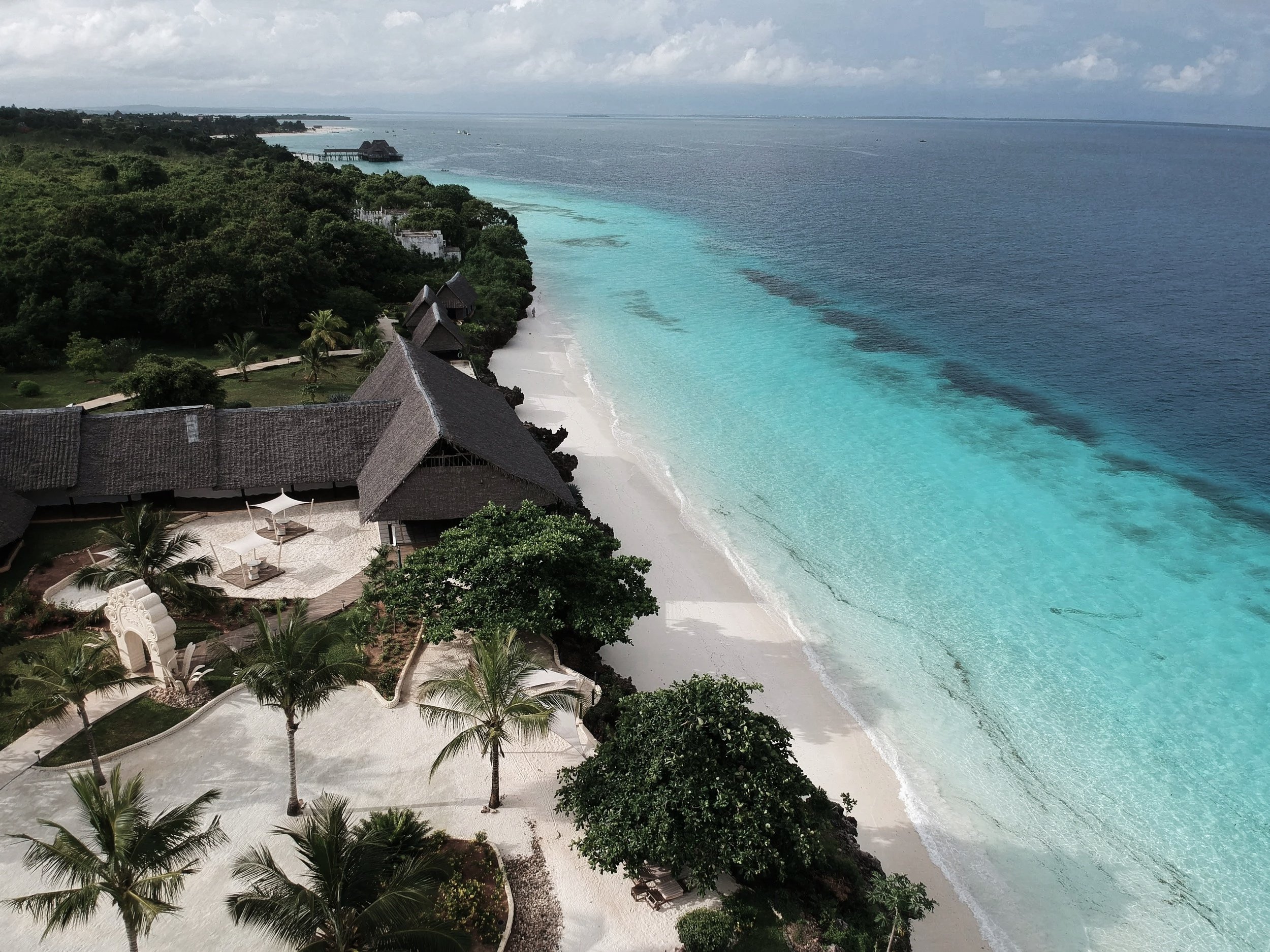 Aerial view of a tropical beach resort with white sand, turquoise water, palm trees, and thatched-roof buildings on the coast.