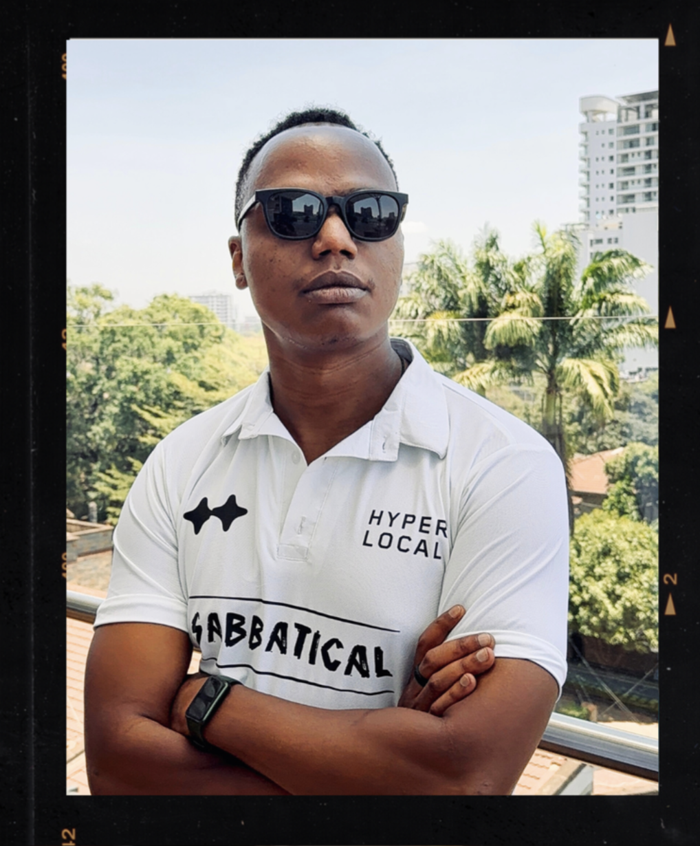 A man in sunglasses wearing a white polo shirt with black patch and text, standing outdoors on a balcony with green trees, palm trees, and buildings in the background.