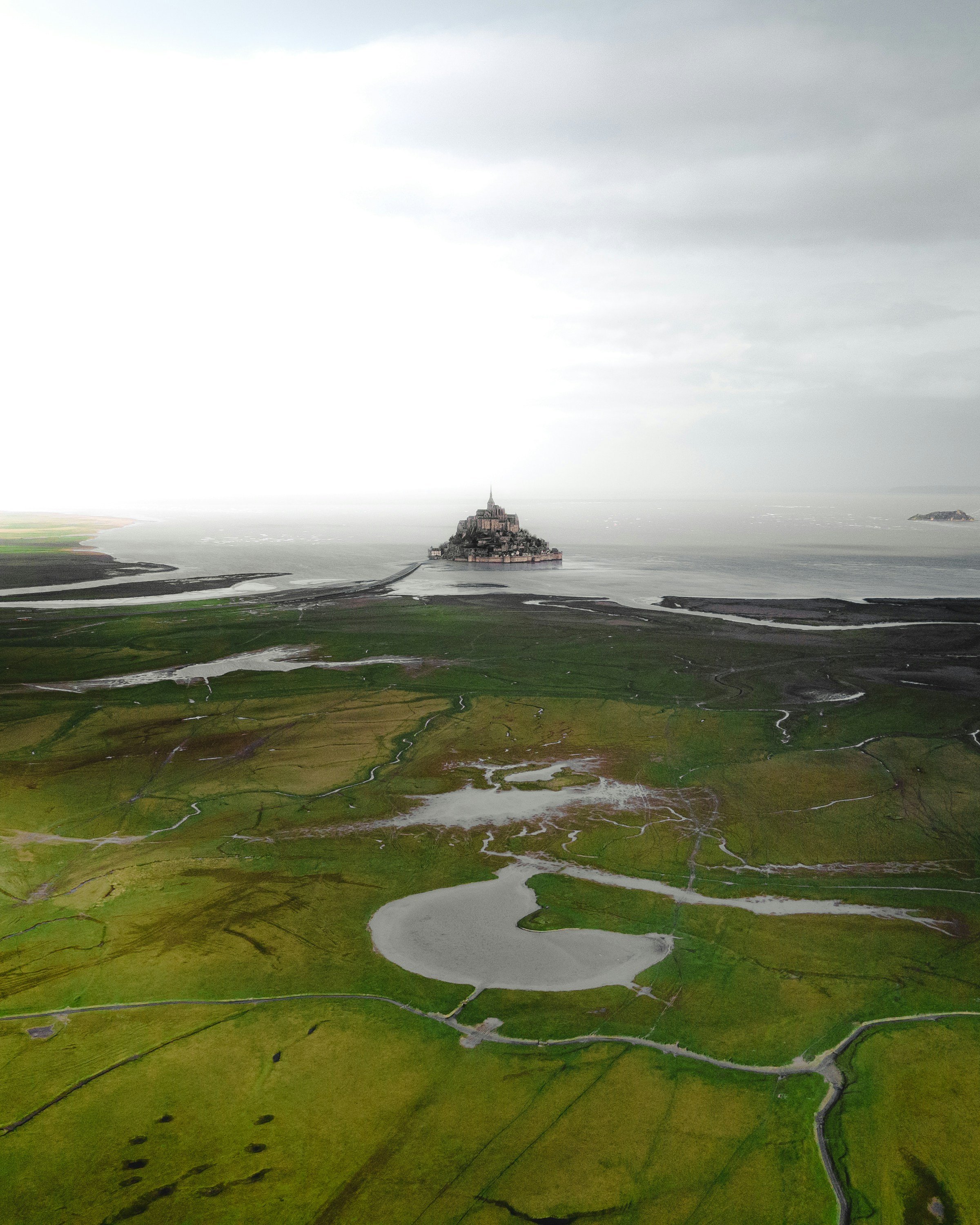 Aerial view of Mont Saint-Michel, a historic island commune in Normandy, France, surrounded by green fields, waters, and connected to the mainland by a causeway under an overcast sky.