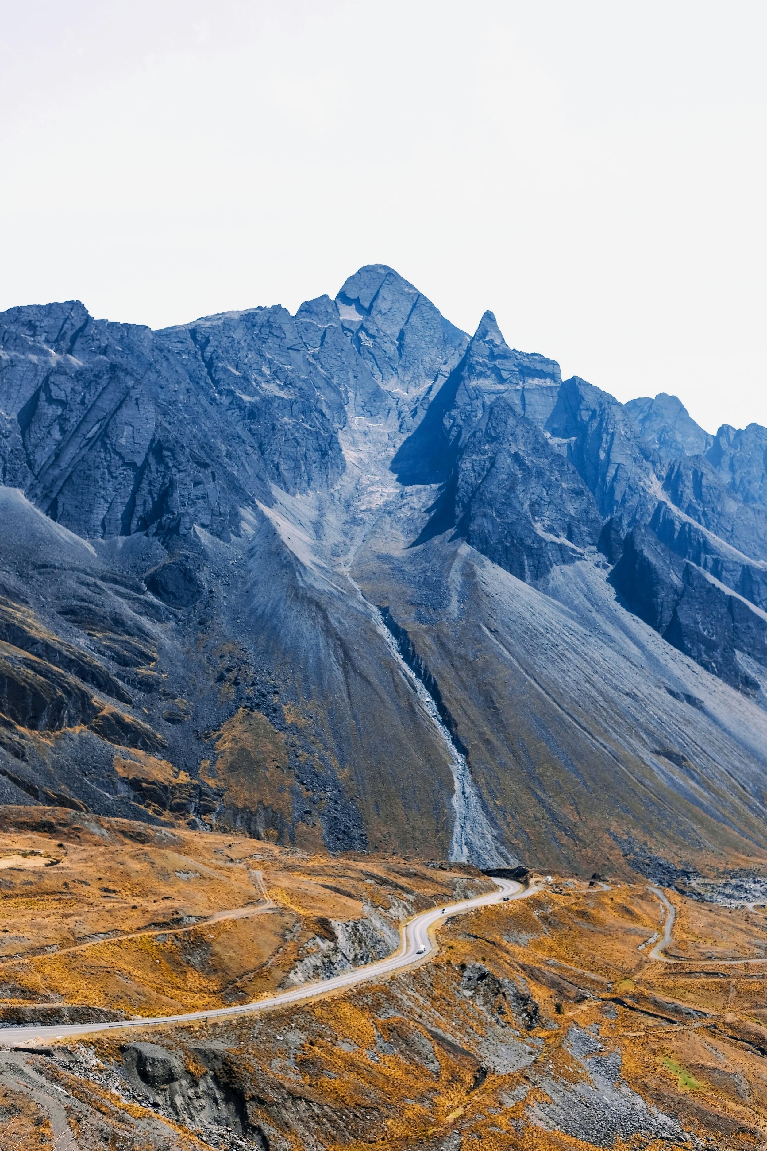 Mountain landscape with rugged peaks, a winding road, and dry, grassy terrain in the foreground.