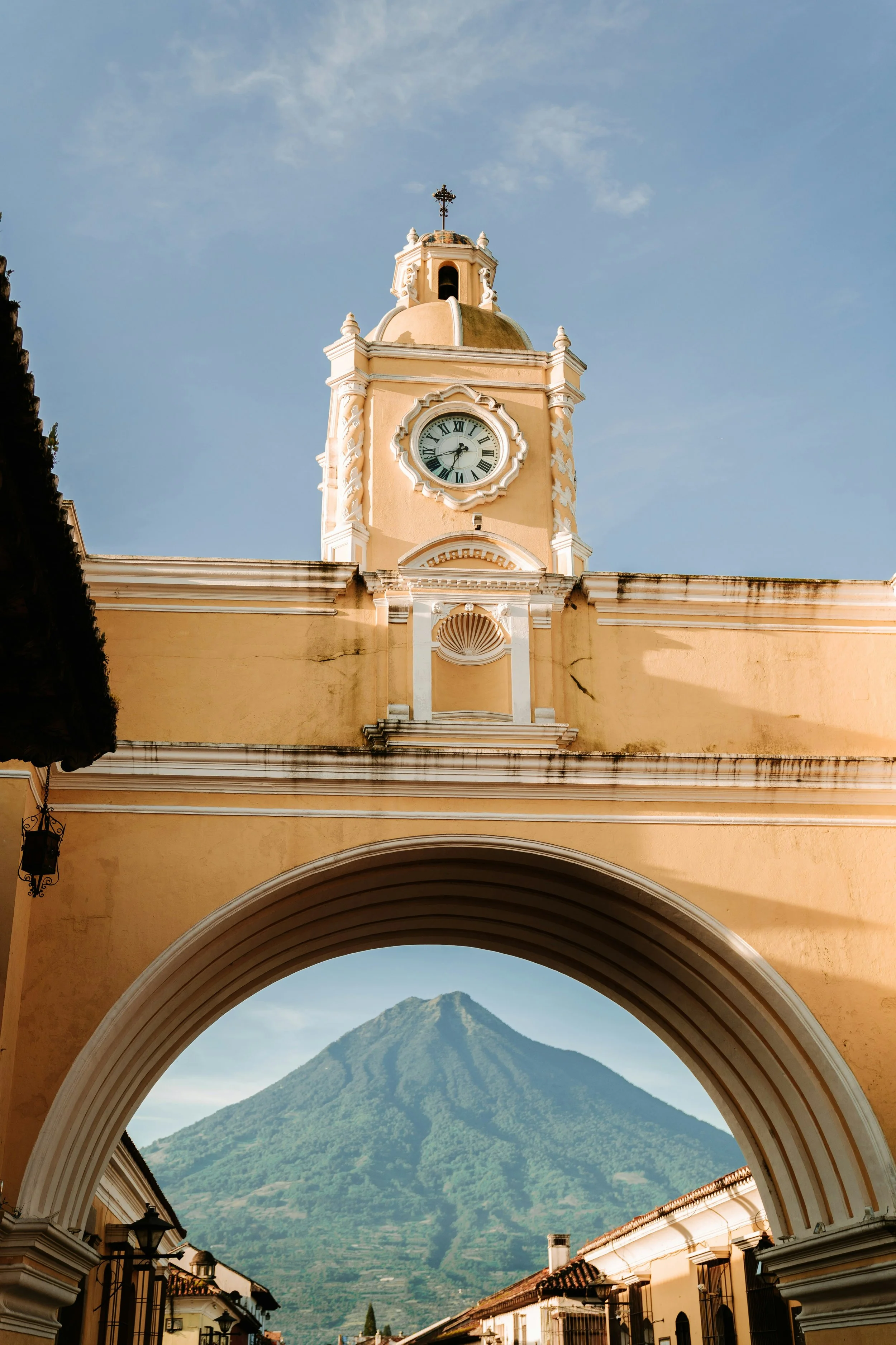 A historic building with a clock tower and a cross on top, arched entrance framing a volcano in the background.