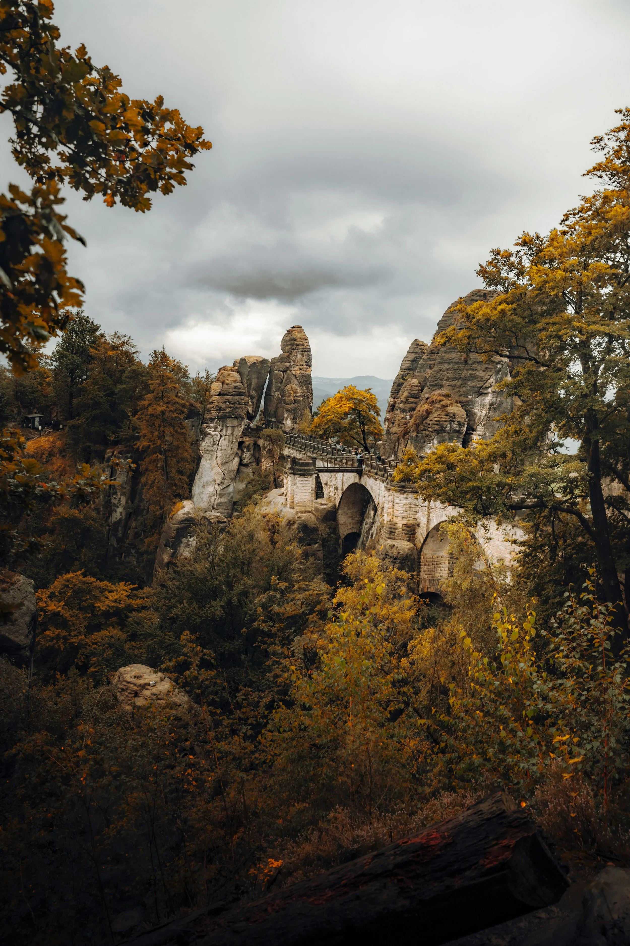Scenic view of a stone bridge crossing a deep valley surrounded by autumn trees and large rock formations under a cloudy sky.
