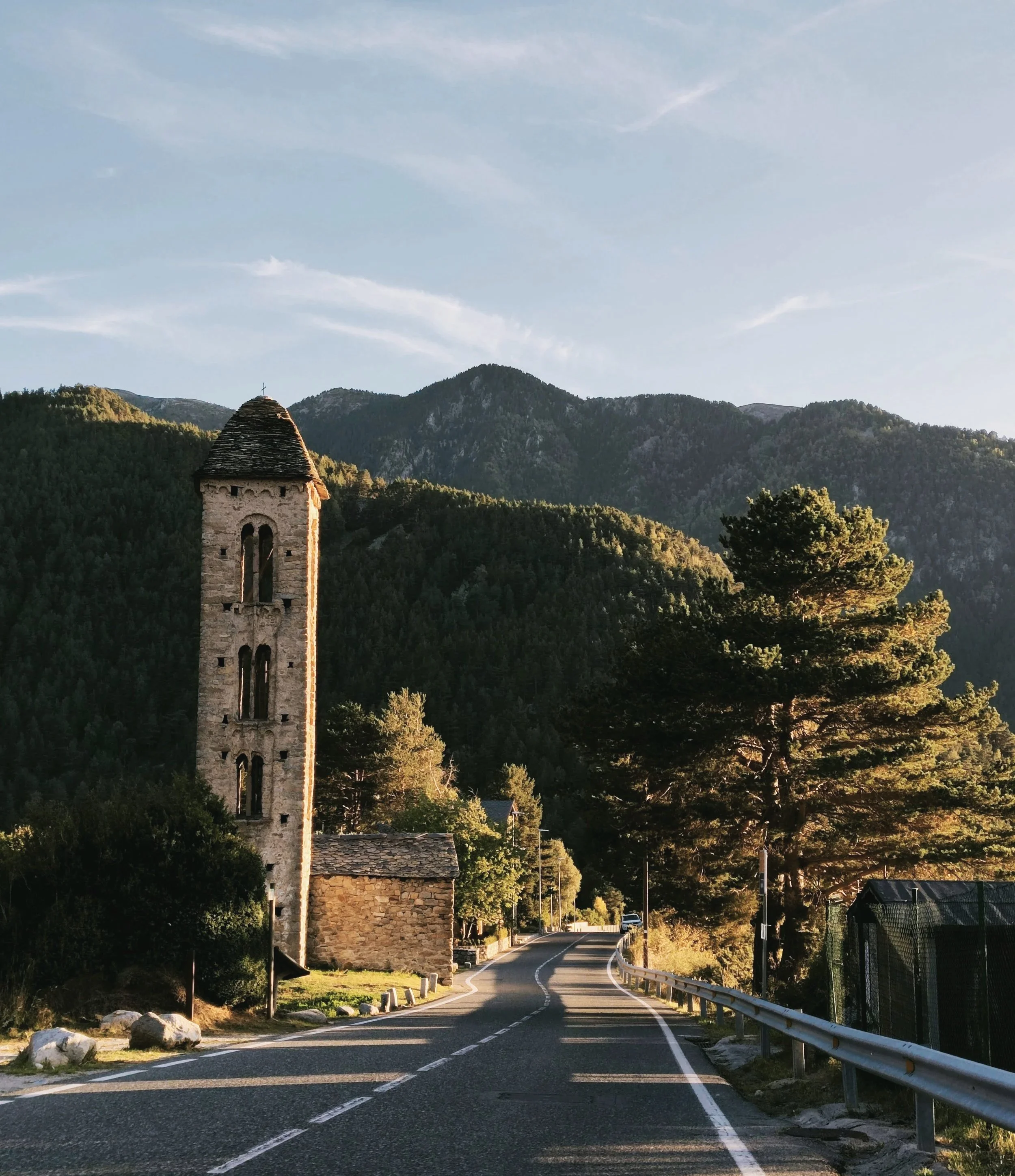 A winding road with a stone tower on the left, surrounded by trees and mountains in the background during sunset.