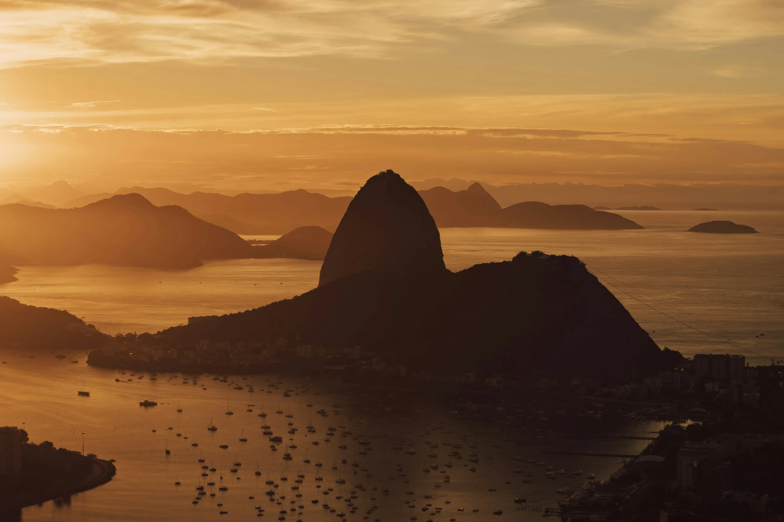 Sunset over Sugarloaf Mountain in Rio de Janeiro, Brazil, with boats in the bay and mountain ranges in the background.