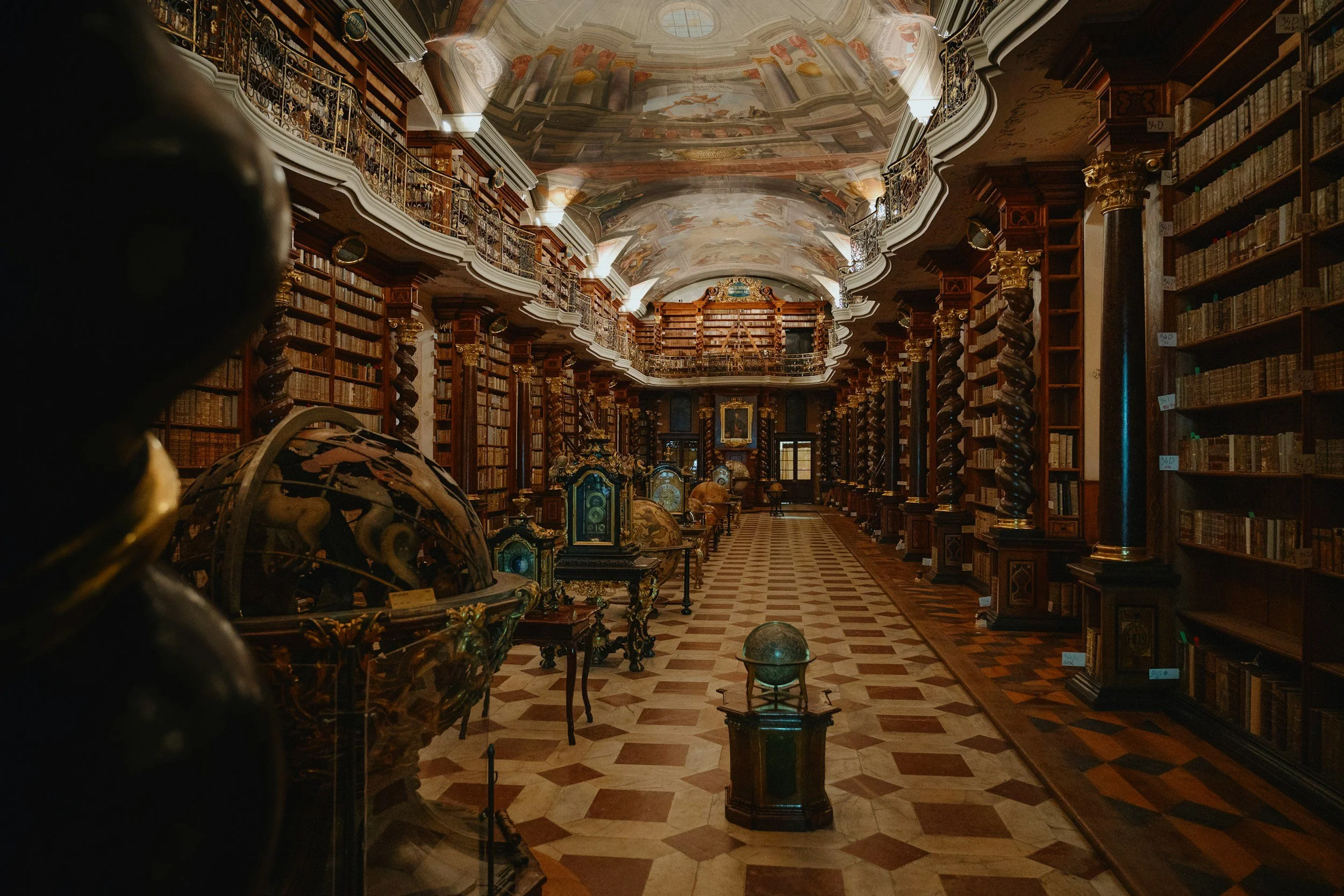 An ornate historic library with tall wooden shelves filled with books, decorative columns, and a painted ceiling. There are globes and antique artifacts displayed along the aisle.