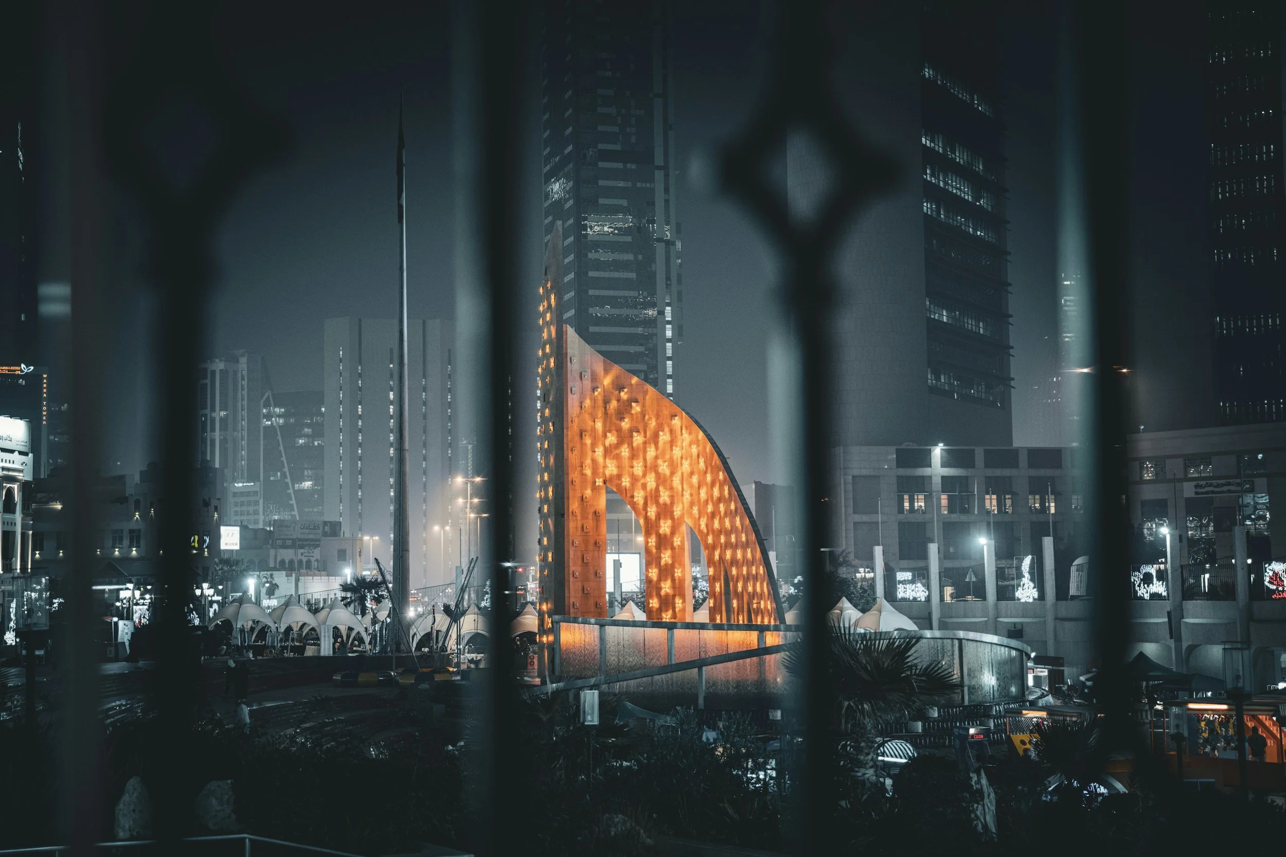 Nighttime cityscape viewed through a fence with the illuminated Dubai Frame in the background.