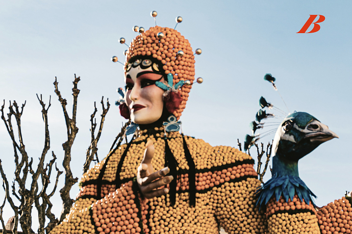 A colorful parade float featuring a person dressed in an elaborate, vibrant costume with beaded headgear, face makeup, and an owl figure, set against a clear sky and leafless trees.