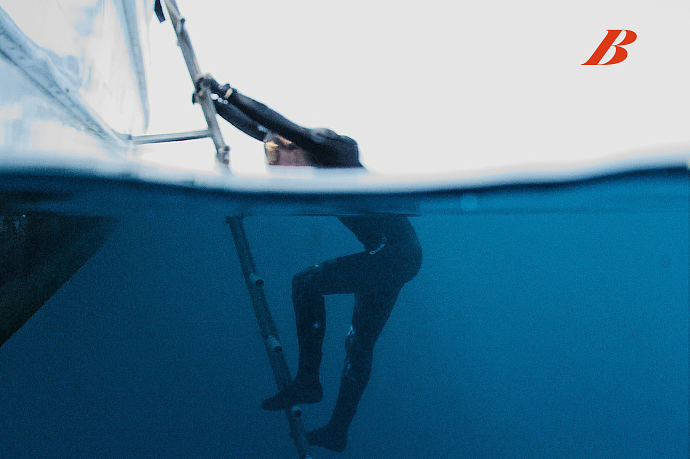 Person climbing a ladder from underwater to above water on a boat, with a partial red 'B' logo in the top right corner.