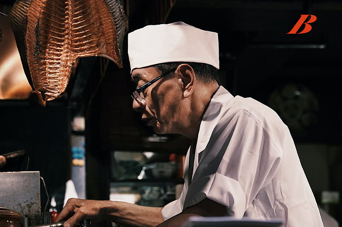 Chef in white uniform and hat preparing food in a kitchen, with a large piece of fish or meat hanging overhead.