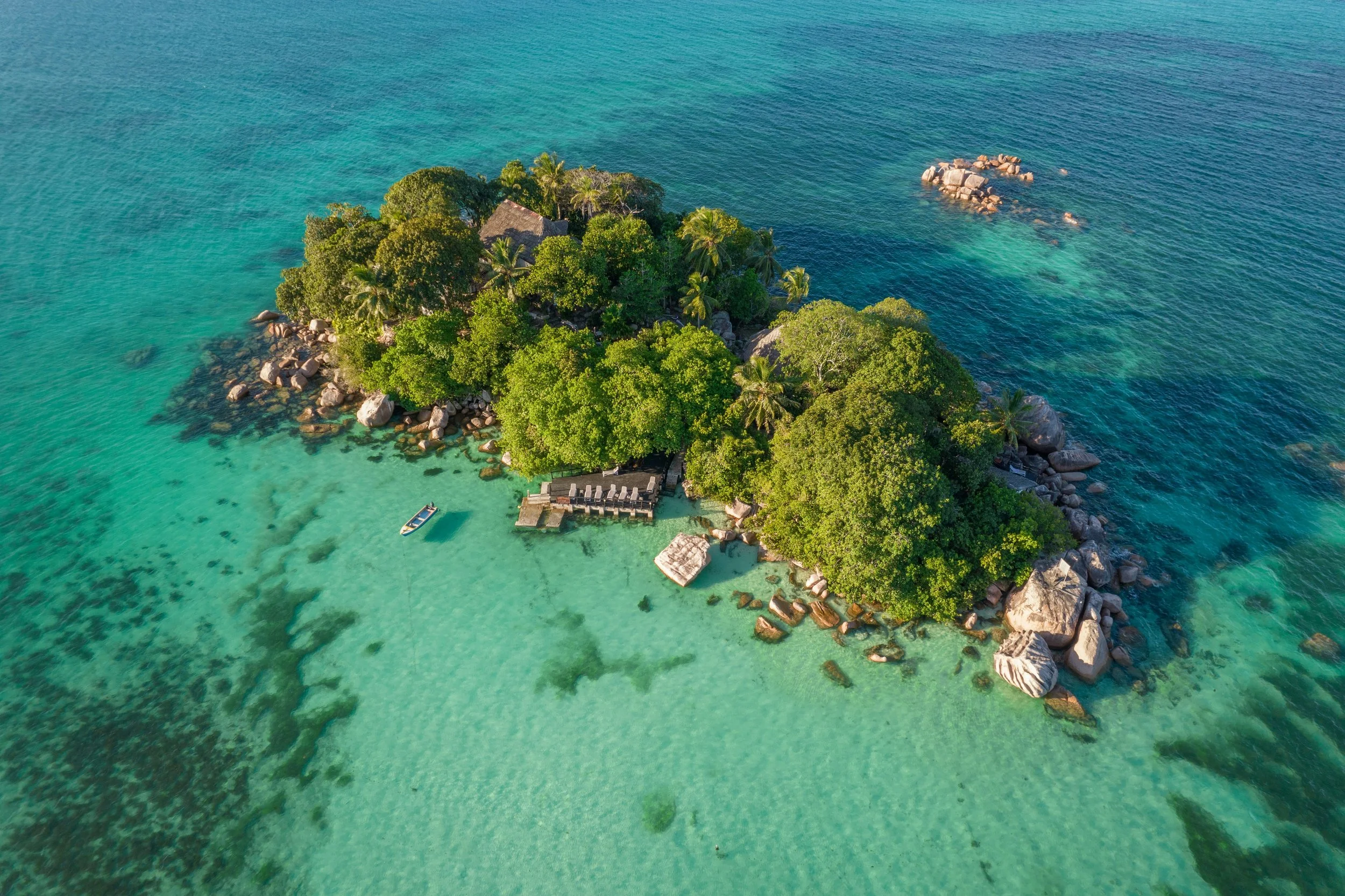 Aerial view of a small tropical island surrounded by turquoise waters, with lush green trees, rocks along the shoreline, and a small boat anchored near a wooden dock.