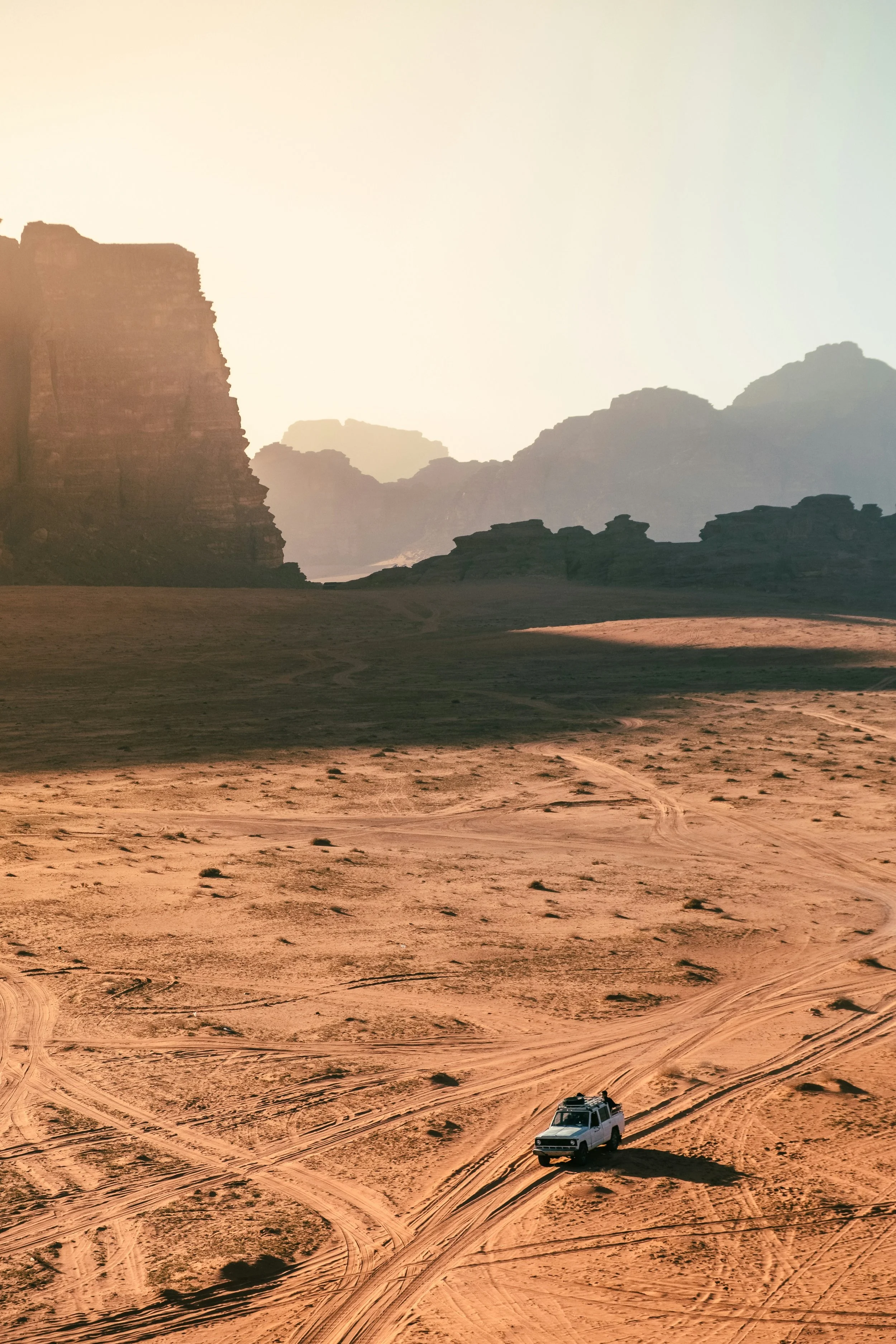 A white SUV driving on a sandy desert with tire tracks, with rocky cliffs and mountains in the background under a bright sky.