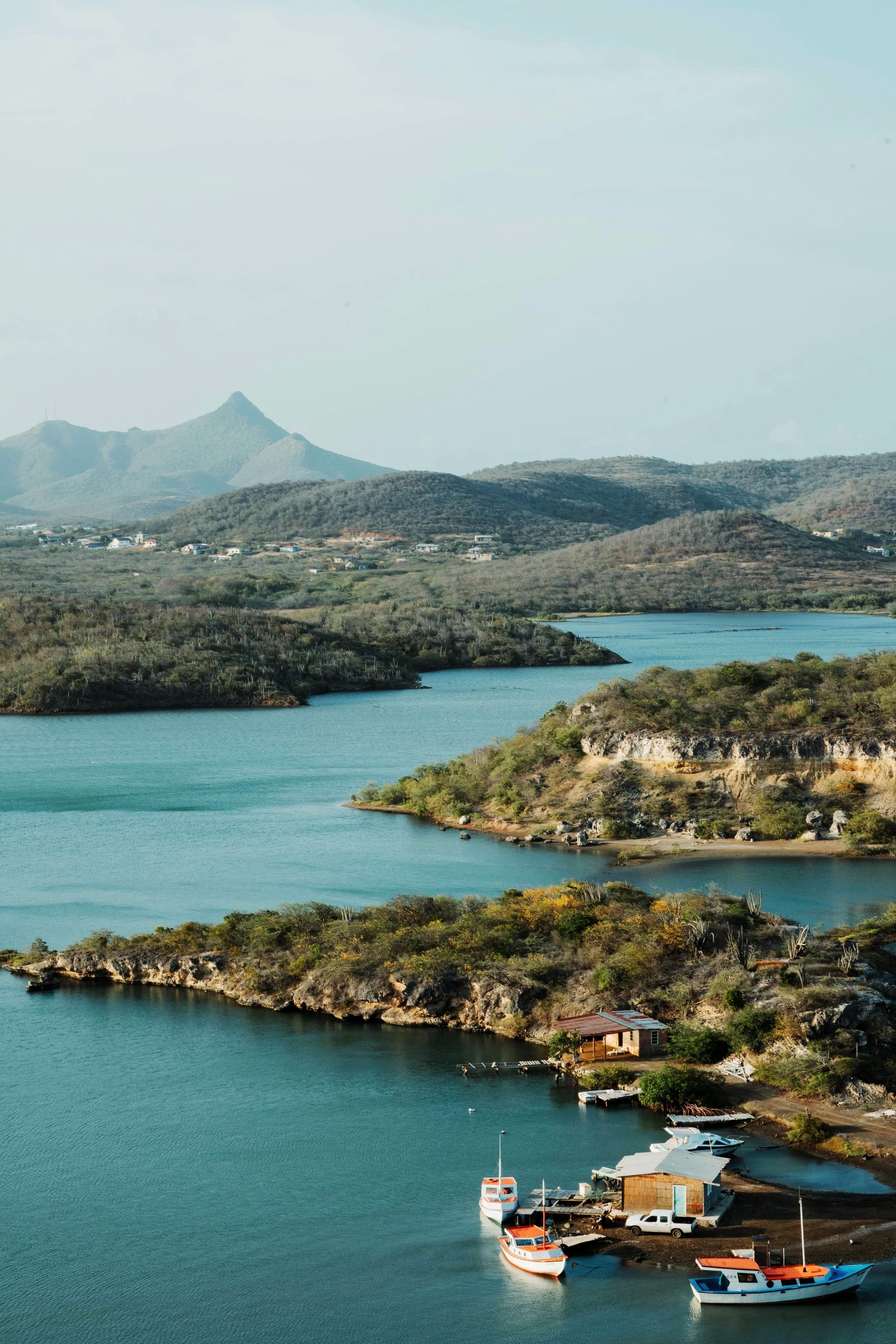 A scenic view of a large lake with several boats docked near a small wooden pier, surrounded by rolling hills and distant mountains.