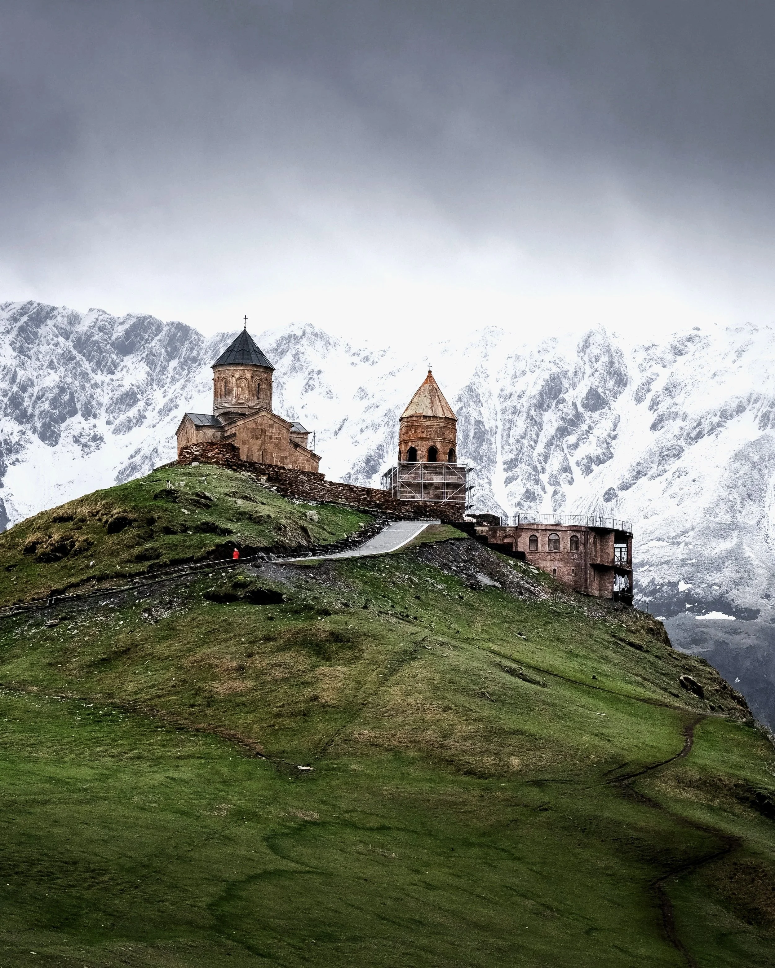 A stone church with two towers on a grassy hill, with snow-capped mountains in the background under a cloudy sky.