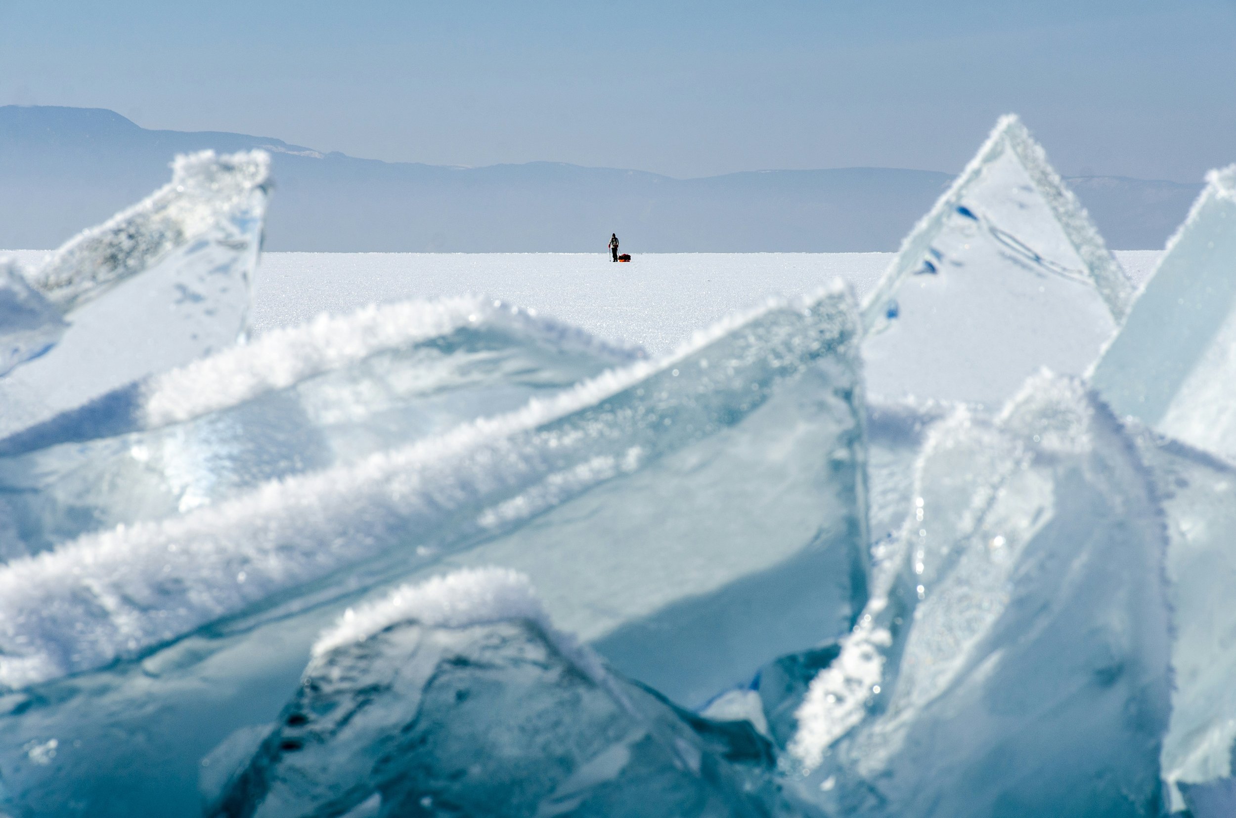 A person wearing winter clothing with a sled on a vast, frozen, snow-covered landscape with large chunks of icy ice in the foreground and mountains in the background.
