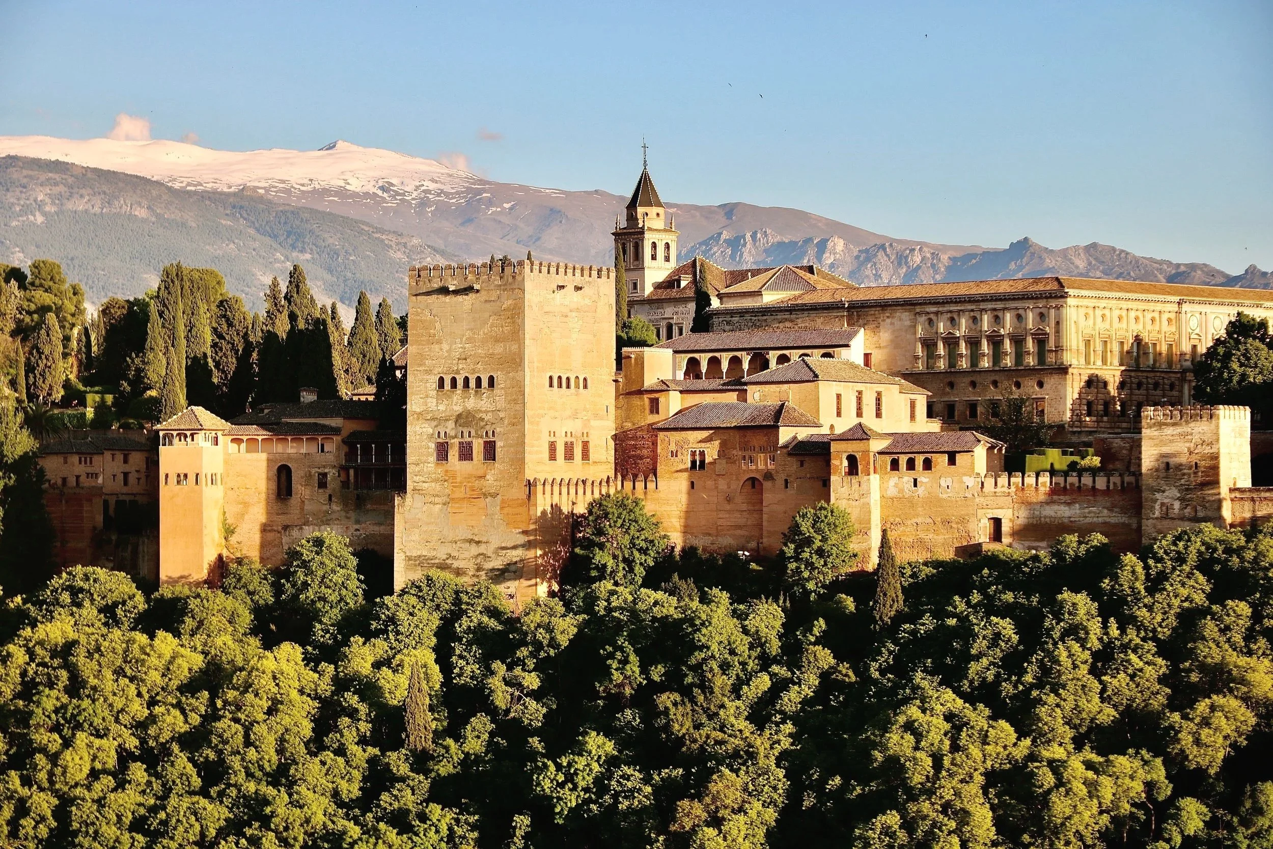 Alhambra palace and fortress complex in Granada, Spain, with snow-capped mountains in the background and green trees in the foreground.
