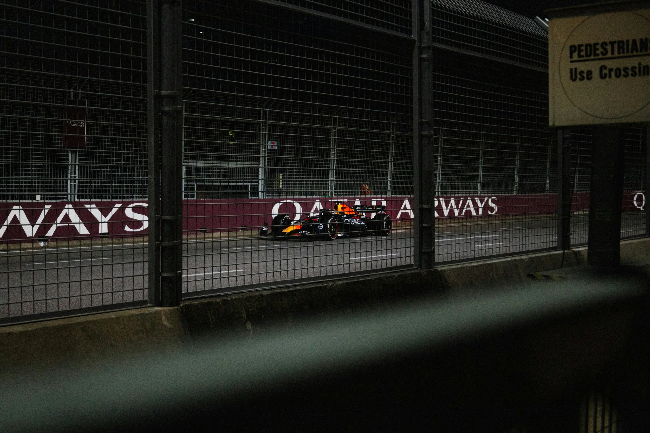 Nighttime scene at a Formula 1 race track with a single race car on the track, visible through a wire fence barrier. The car is painted in dark colors with orange accents. There are advertising banners along the side of the track.