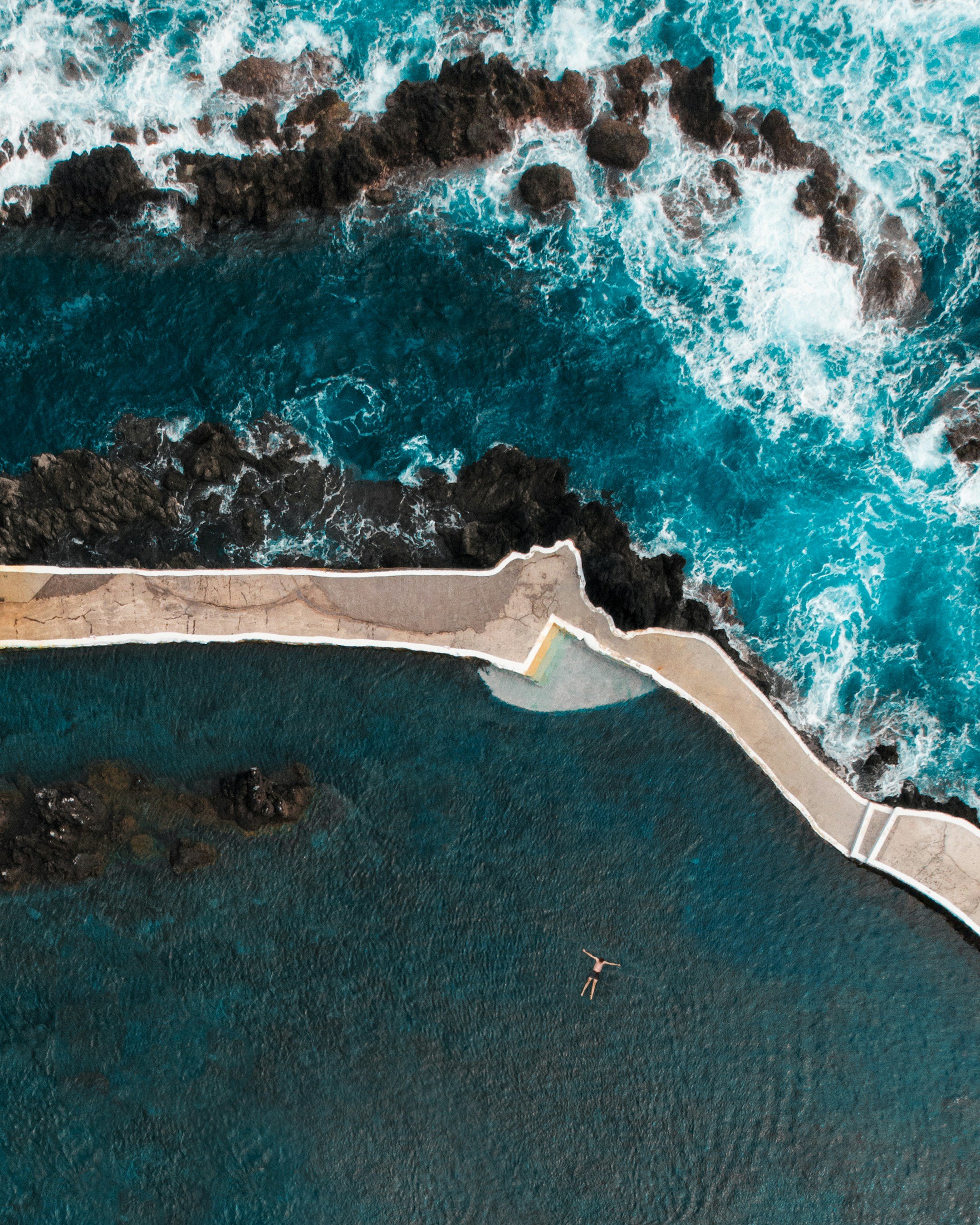 An aerial view of a coastline with a concrete pathway running along a rugged rocky shore. Turquoise ocean waves crash against the rocks, and a person is lying on the water in a swimming pool or calm water area near the pathway.