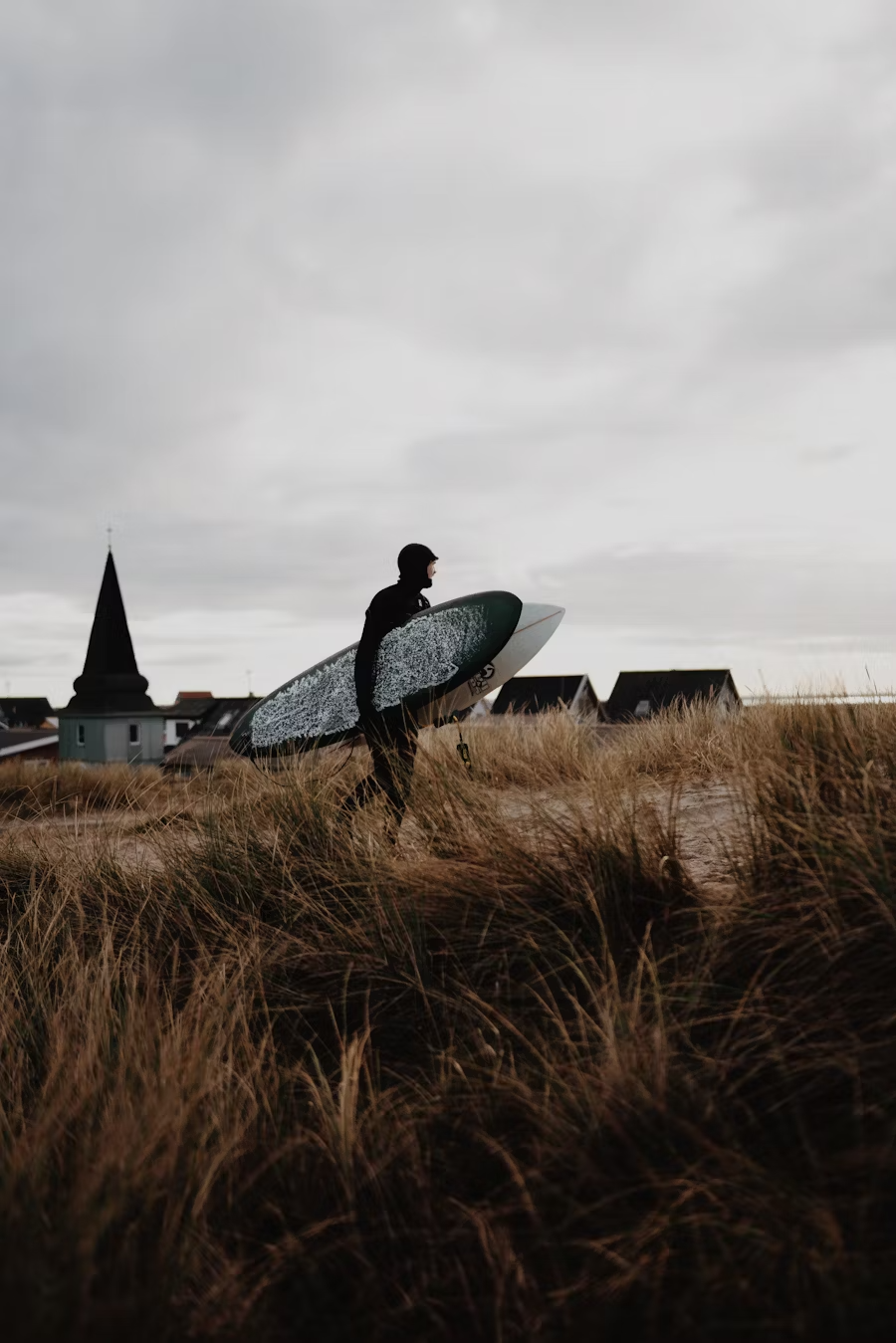Person carrying a surfboard walking through grassy dunes near a coastal town with a church steeple in the background on a cloudy day.