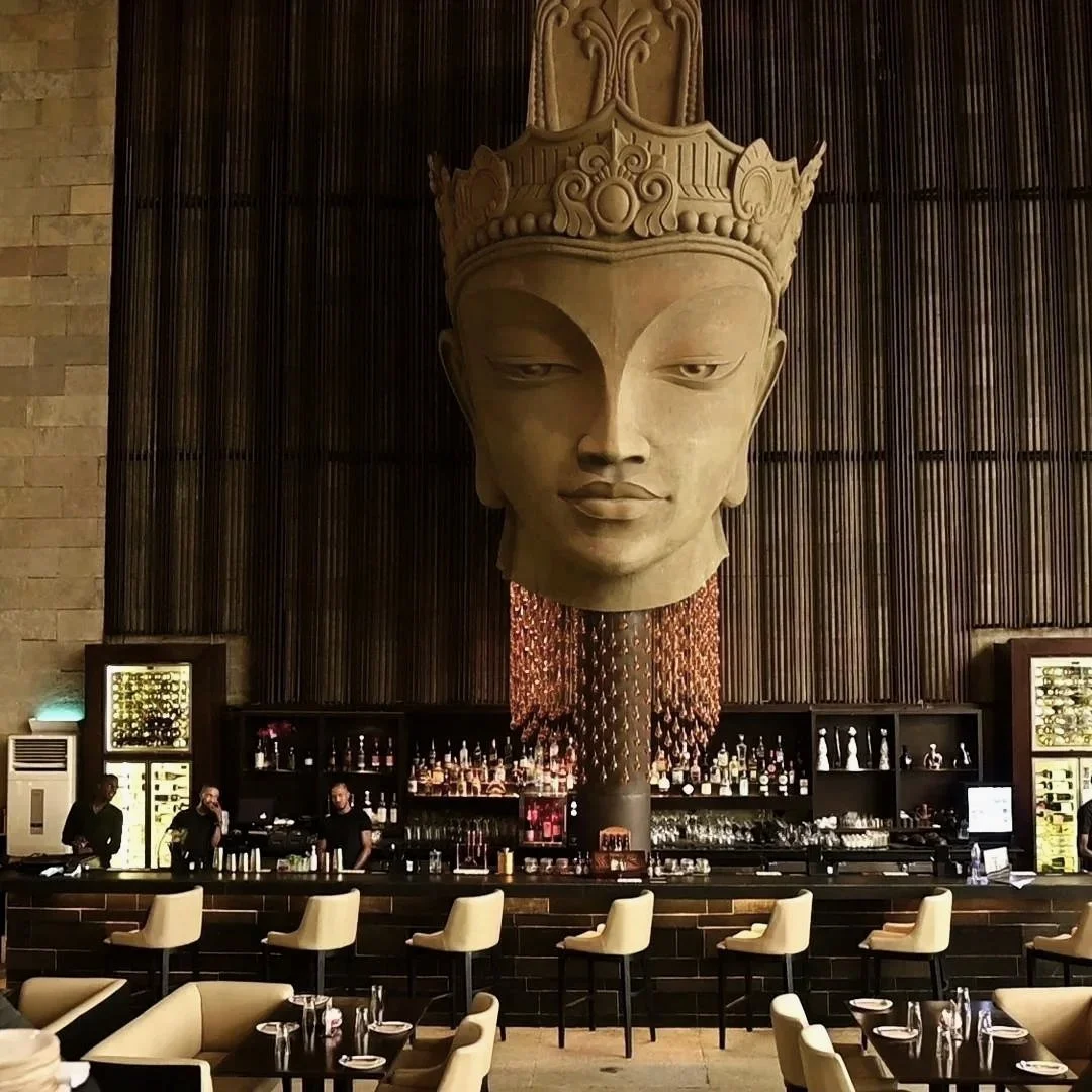 A large decorative sculpture of an Asian woman's face with a crown, hanging above a bar in a sophisticated restaurant or lounge, with bartenders behind the counter and tables with chairs in the foreground.