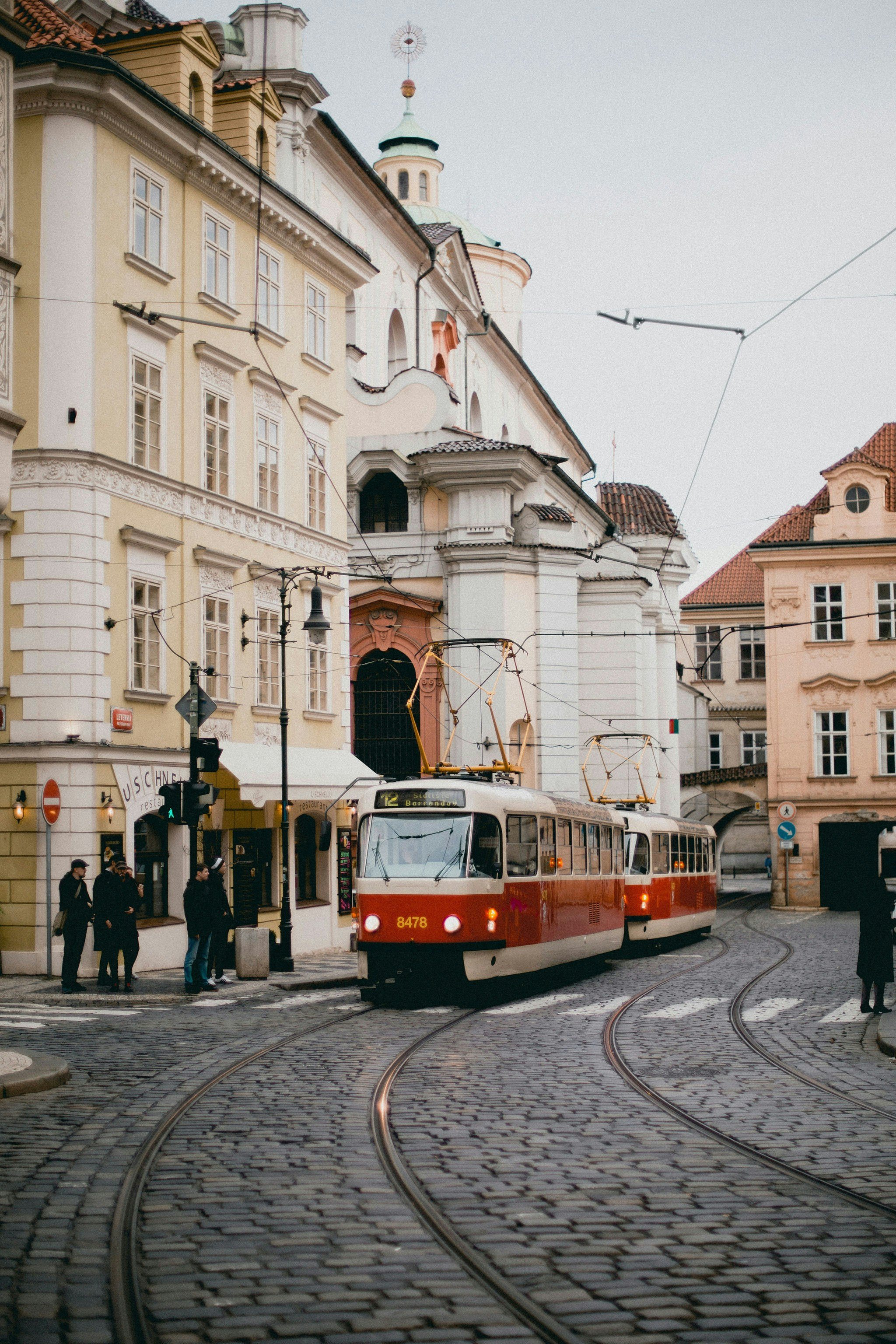 A red and white tram moving through a historic European city street with cobblestone pavement, surrounded by colorful old buildings and pedestrians, during daytime.
