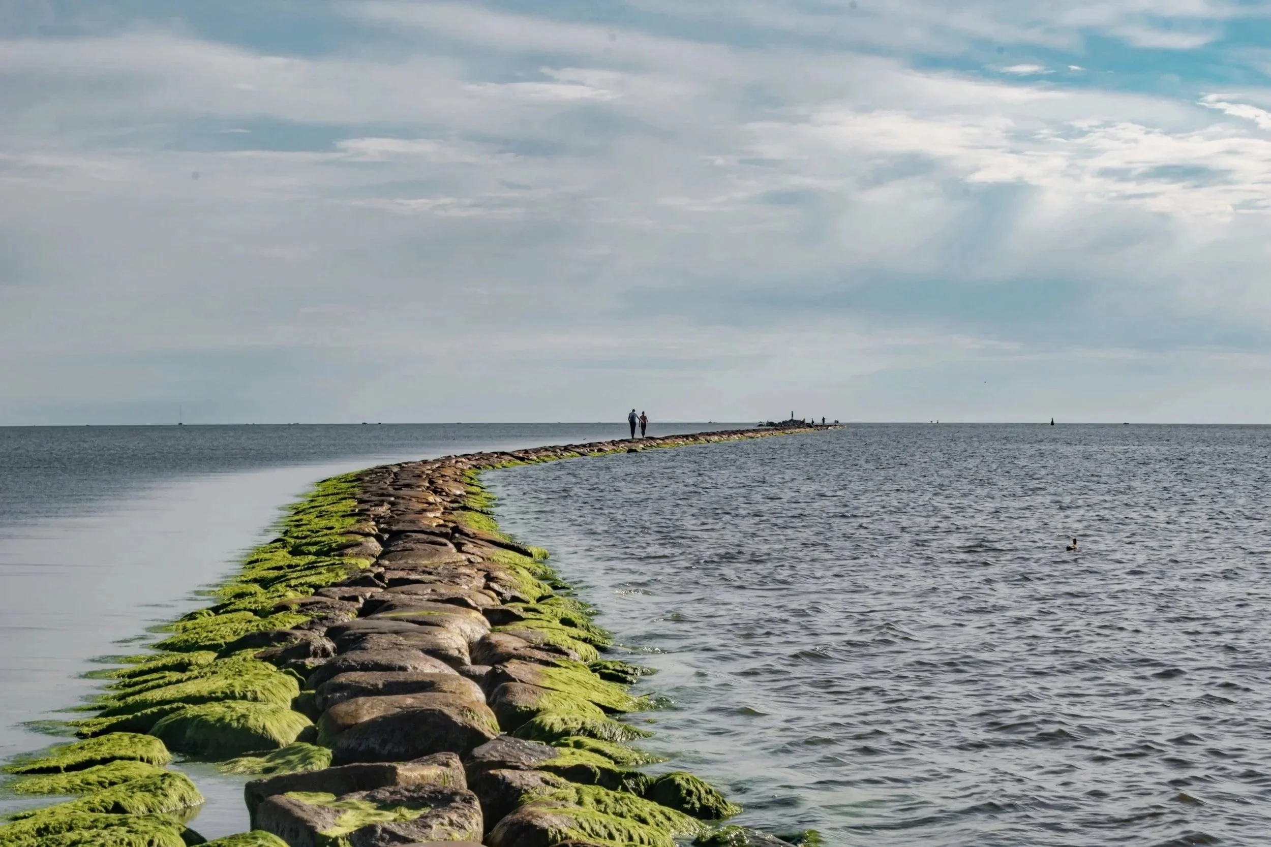 A stone breakwater covered with green moss extending into the water, with two people walking at the end of it, under a cloudy sky.