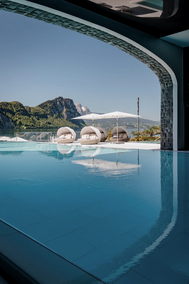 View of an indoor pool with an open wall showing a scenic outdoor landscape with mountains and water, featuring white lounge chairs and umbrellas.