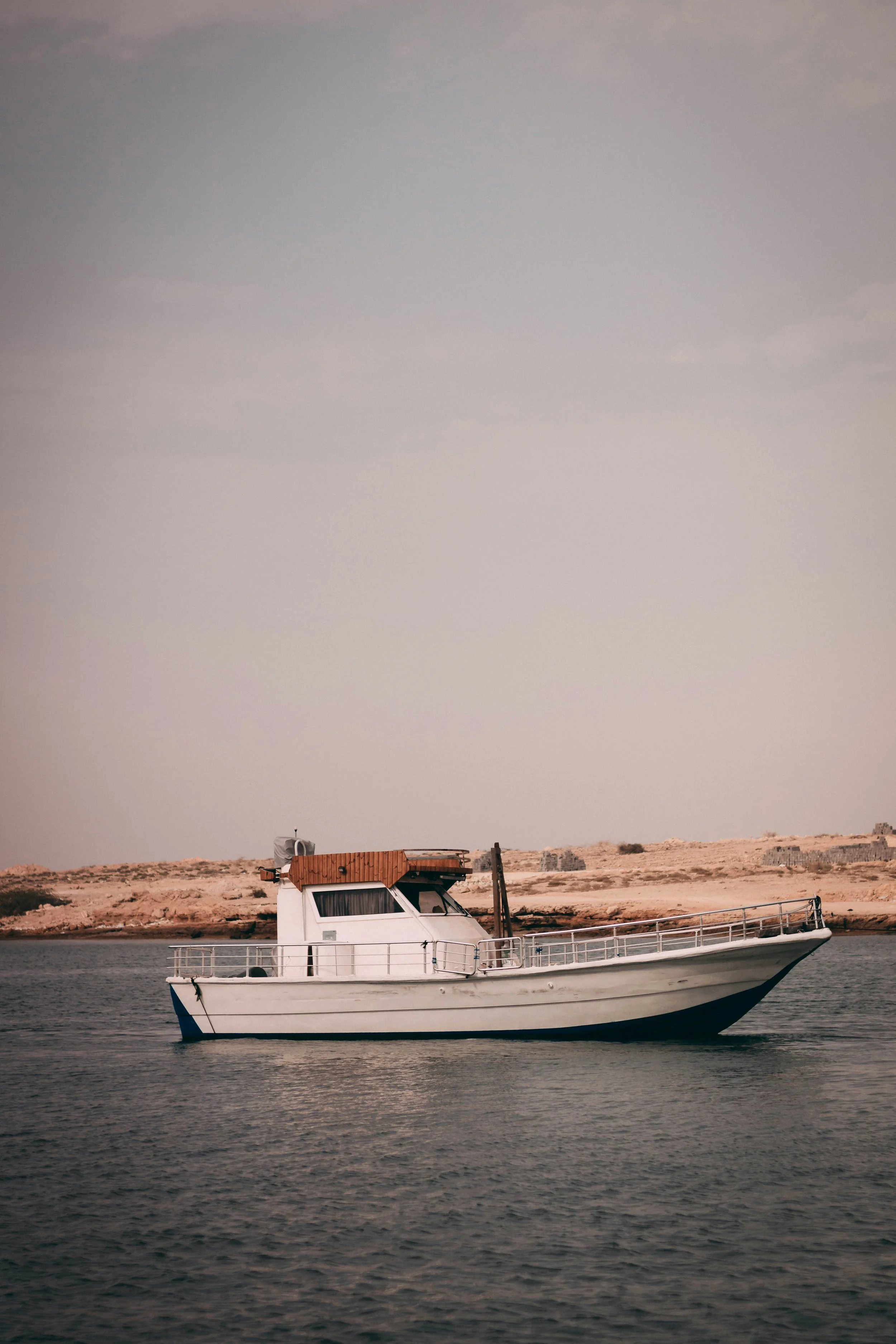 A white boat with a wooden cabin on a body of water, near a rocky shoreline with minimal vegetation and an overcast sky.