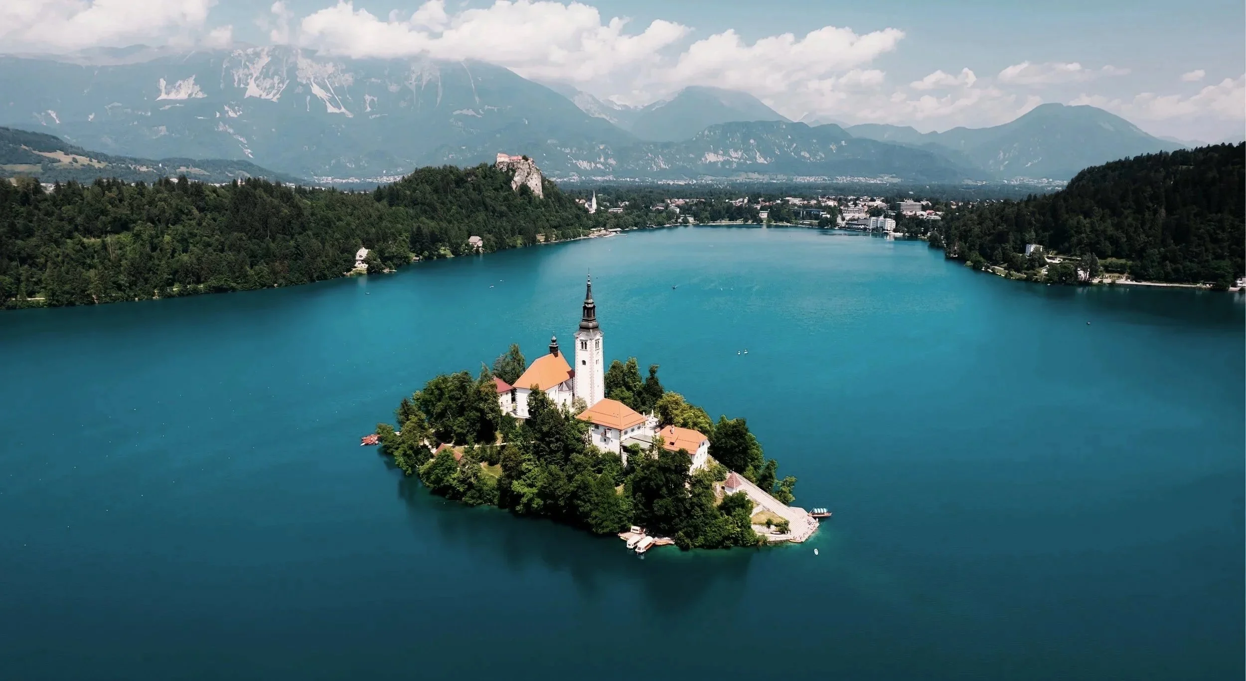 Aerial view of Lake Bled in Slovenia, featuring a small island with a church and a steeple surrounded by calm turquoise waters, with forested hills and mountains in the background.