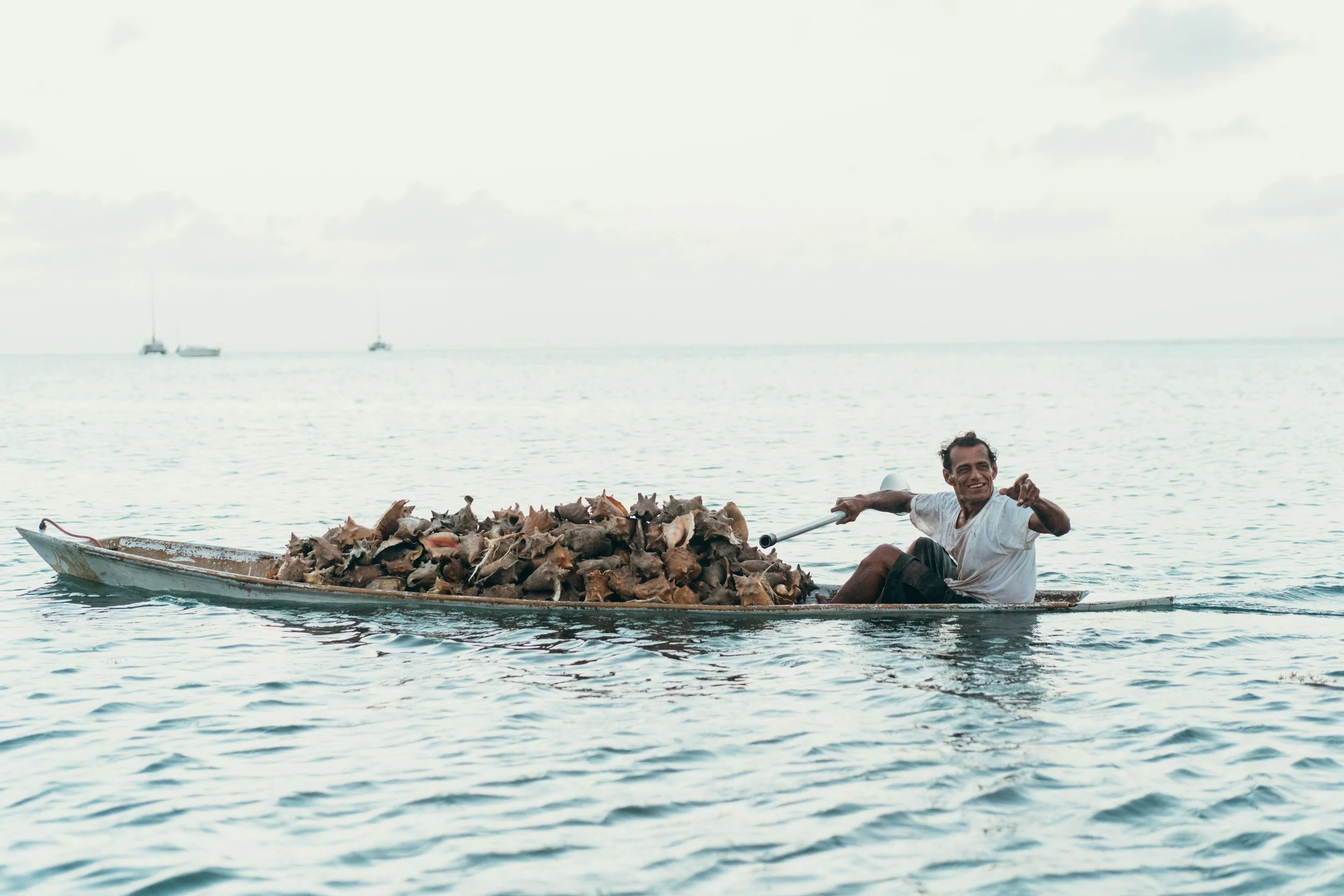 A man paddling a small boat filled with numerous animal skulls and bones in open water with sailboats in the background.