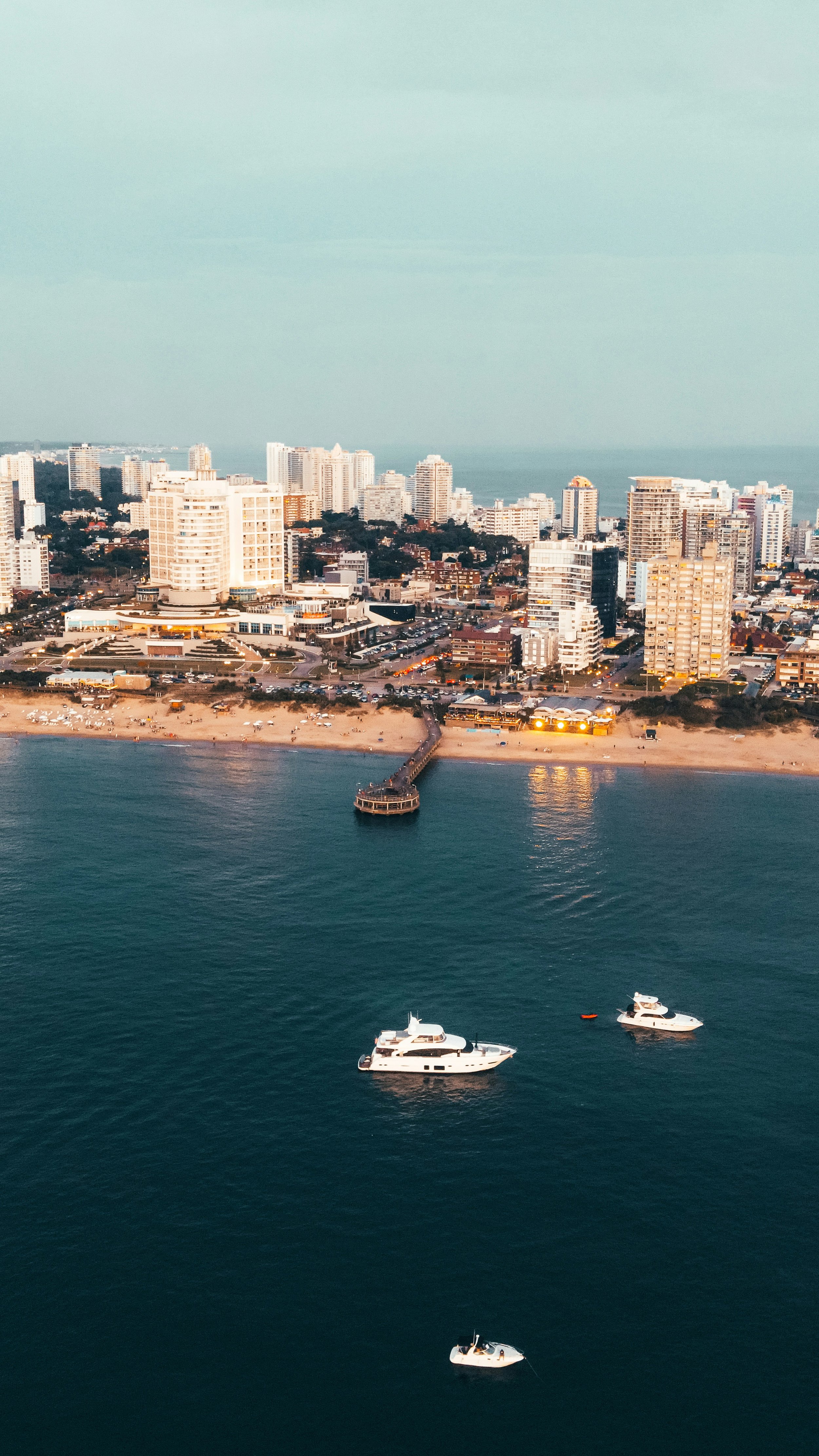 Aerial view of a city skyline with high-rise buildings along a beach, boats in the water, and a pier extending into the sea.