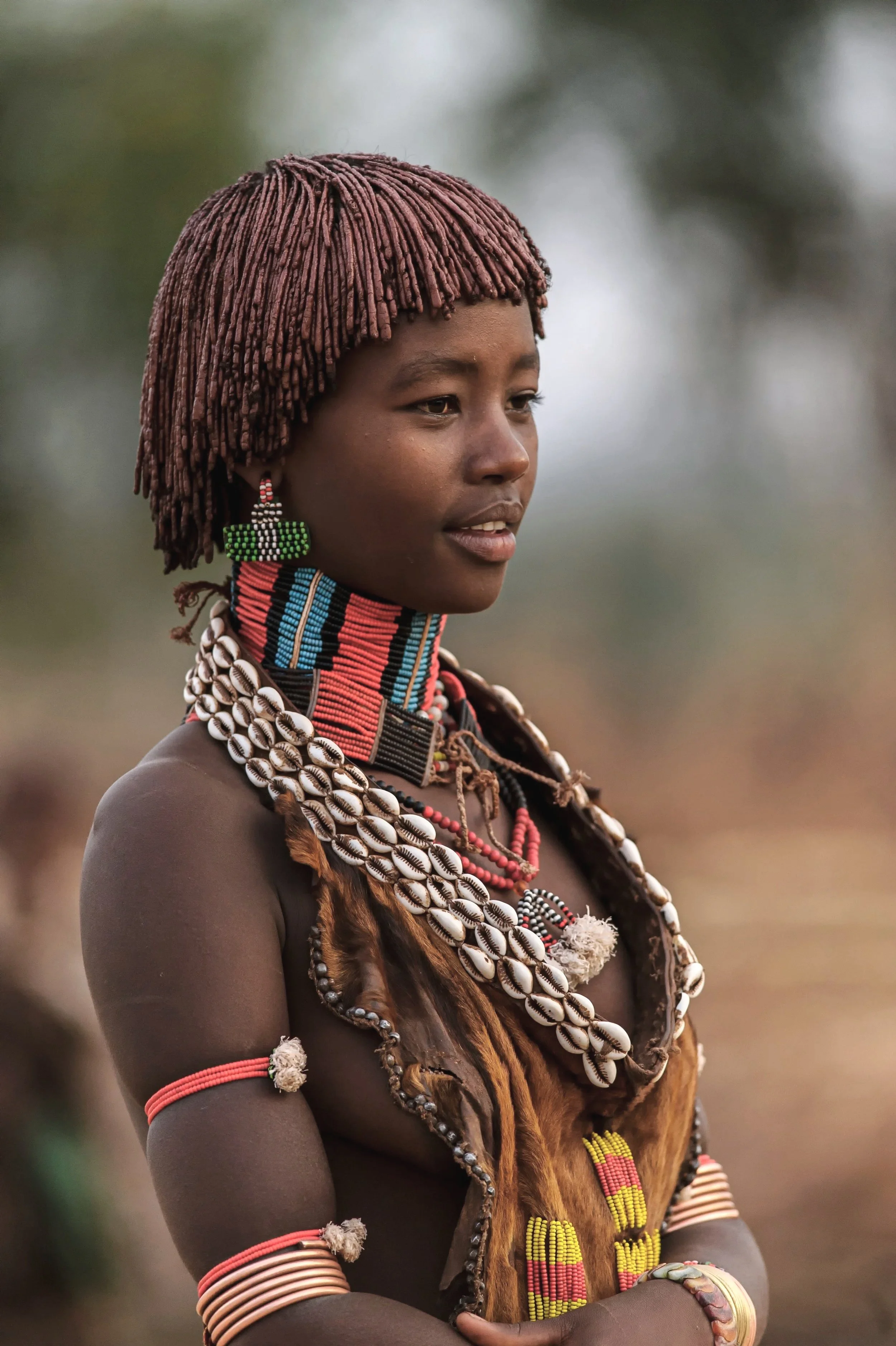 A young woman from an African tribe, wearing traditional beaded jewelry, cowrie shell necklaces, and a fur garment, with her arms crossed.