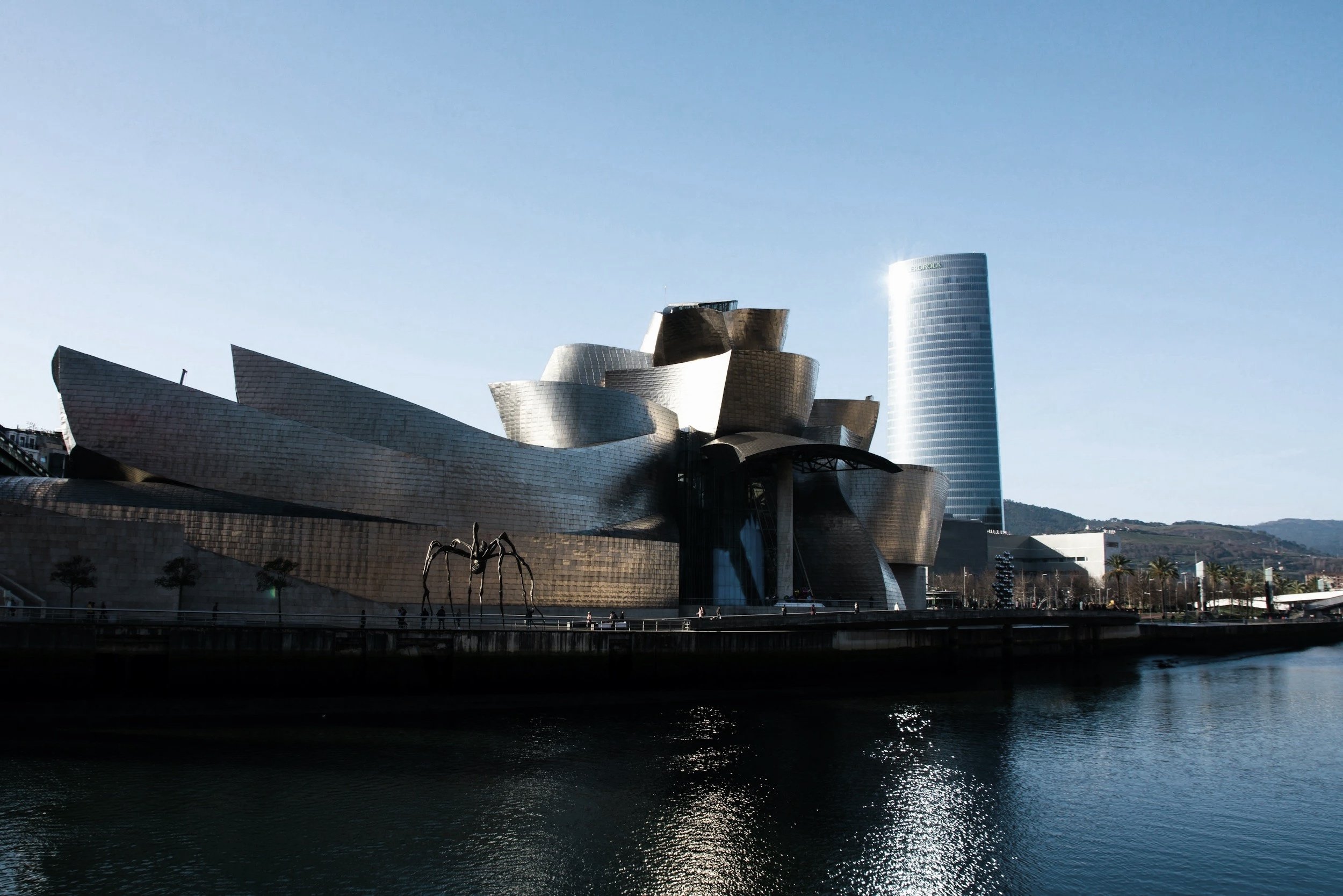 View of the Museo Guggenheim Bilbao with a spider sculpture in the foreground and a modern skyscraper in the background, near a body of water.